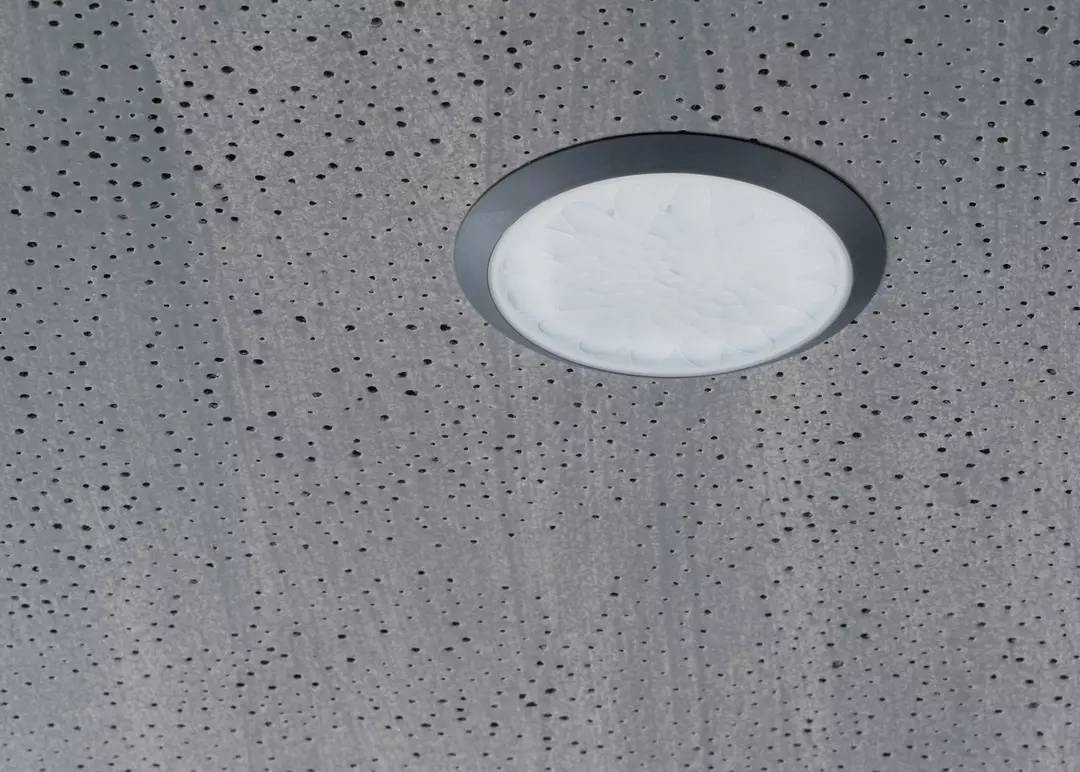 Deckenleuchte in runder Form, eingebettet in eine perforierte, graue Decke. Die Struktur der Decke ist sichtbar.