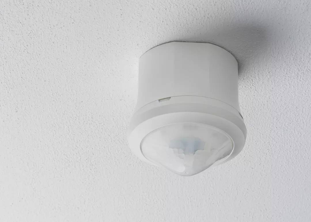 Ein Deckenbewegungsmelder ist an einer weißen Decke montiert. Er dient der automatischen Erkennung von Bewegung zur Steuerung von Beleuchtung oder Alarmanlagen.