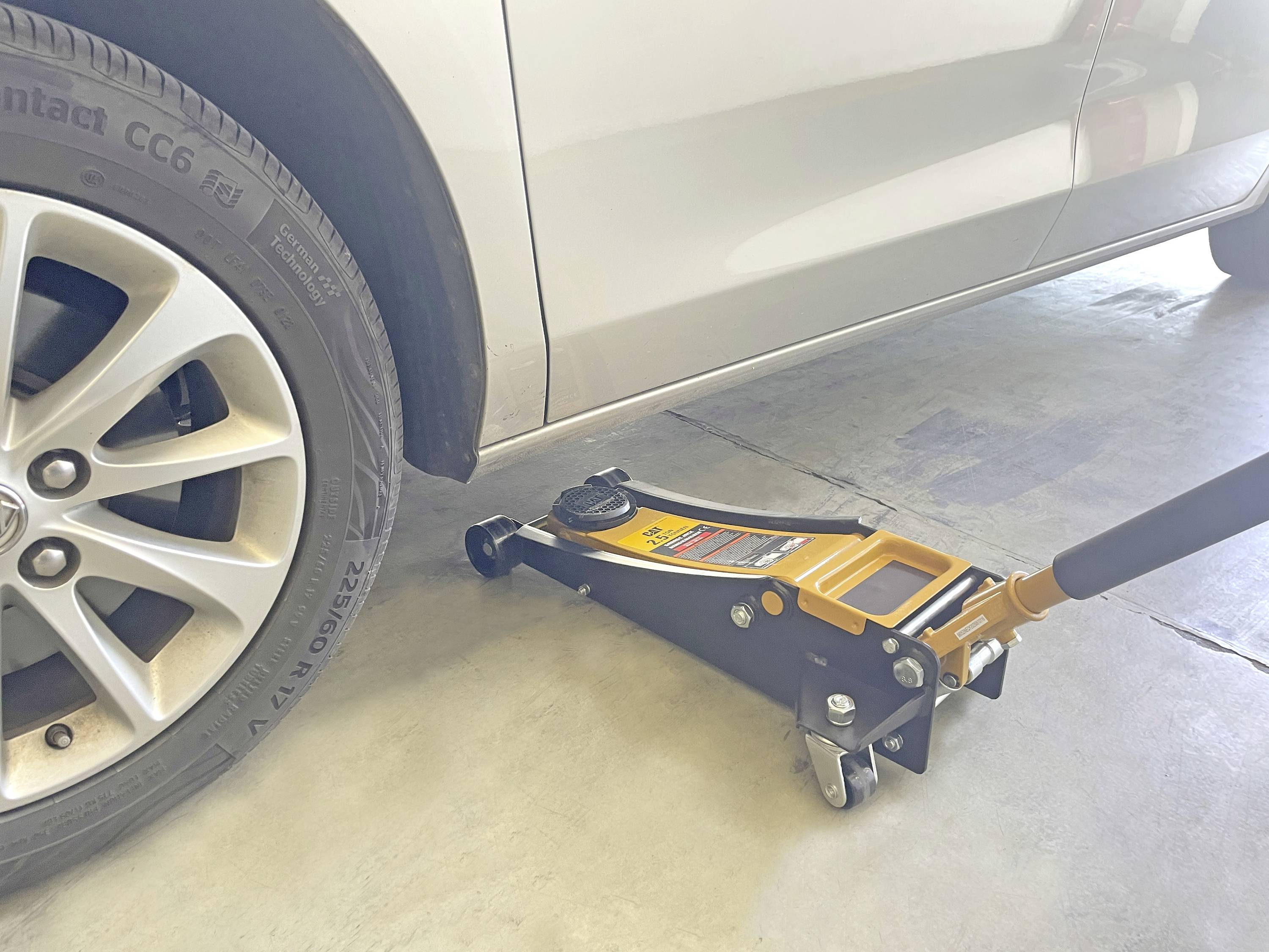 Ein Wagenheber, der ein silbernes Auto in einer Garage hochhebt. Der Wagenheber ist unter dem Rahmen des Fahrzeugs positioniert und hebt das vordere linke Rad vom Boden ab.
