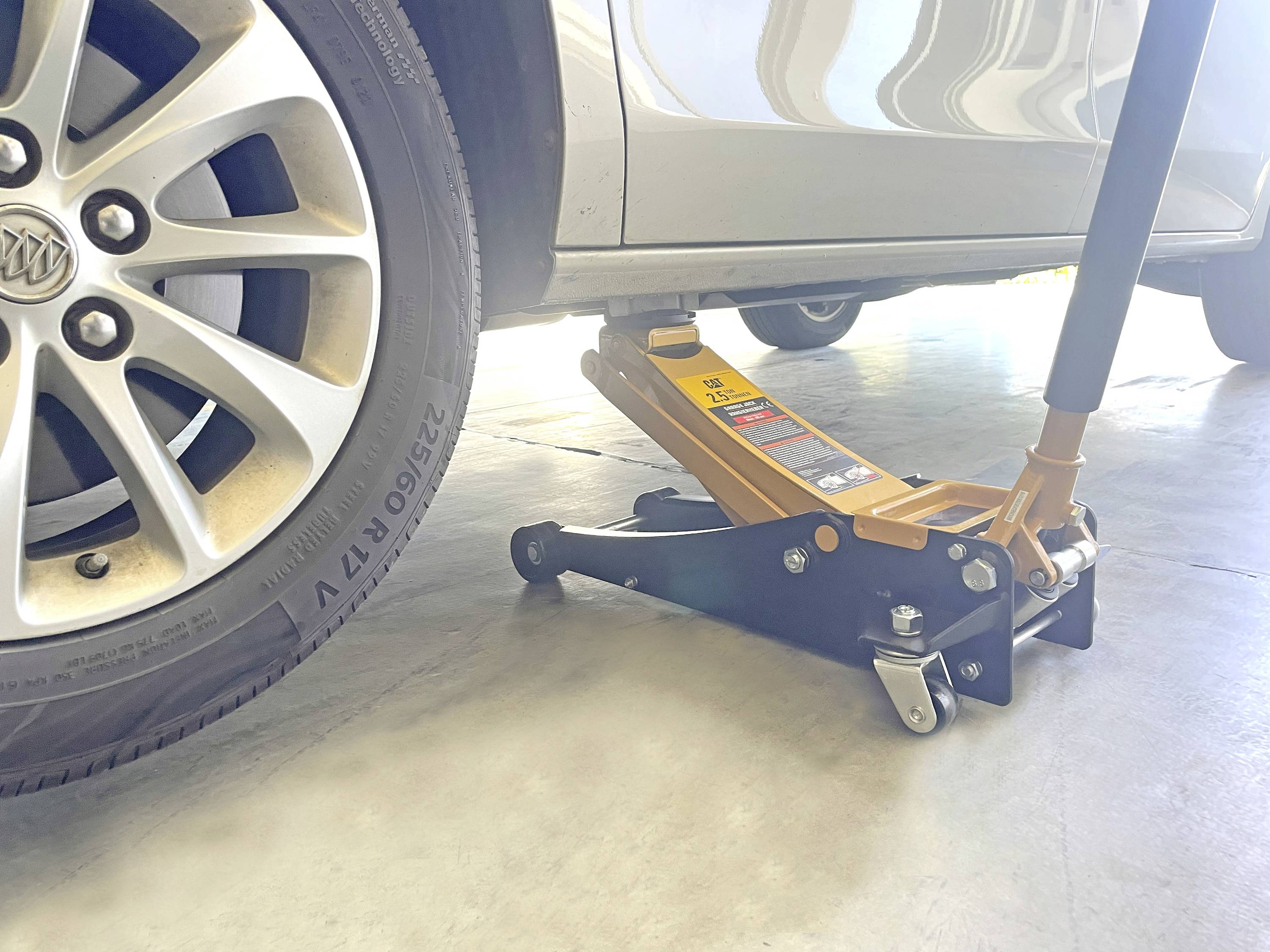 Ein Auto wird in einer Garage mit einem hydraulischen Wagenheber unter dem linken Vorderreifen angehoben. Das Rad des Autos und ein Teil der Karosserie sind sichtbar.