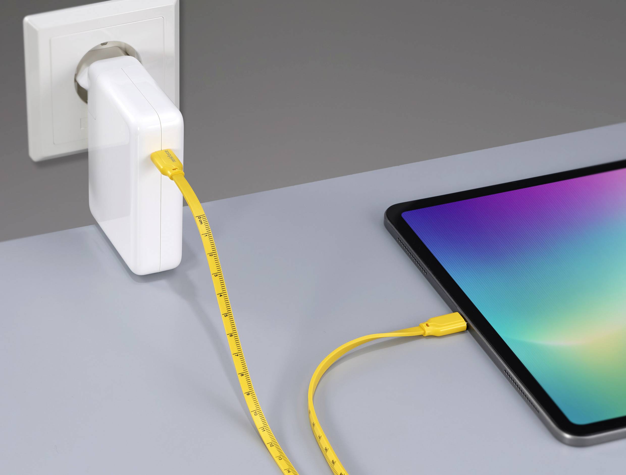 Ein gelbes USB-Kabel verbindet ein Ladegerät, das in einer Steckdose steckt, mit einem Tablet, das auf einem grauen Tisch liegt.
