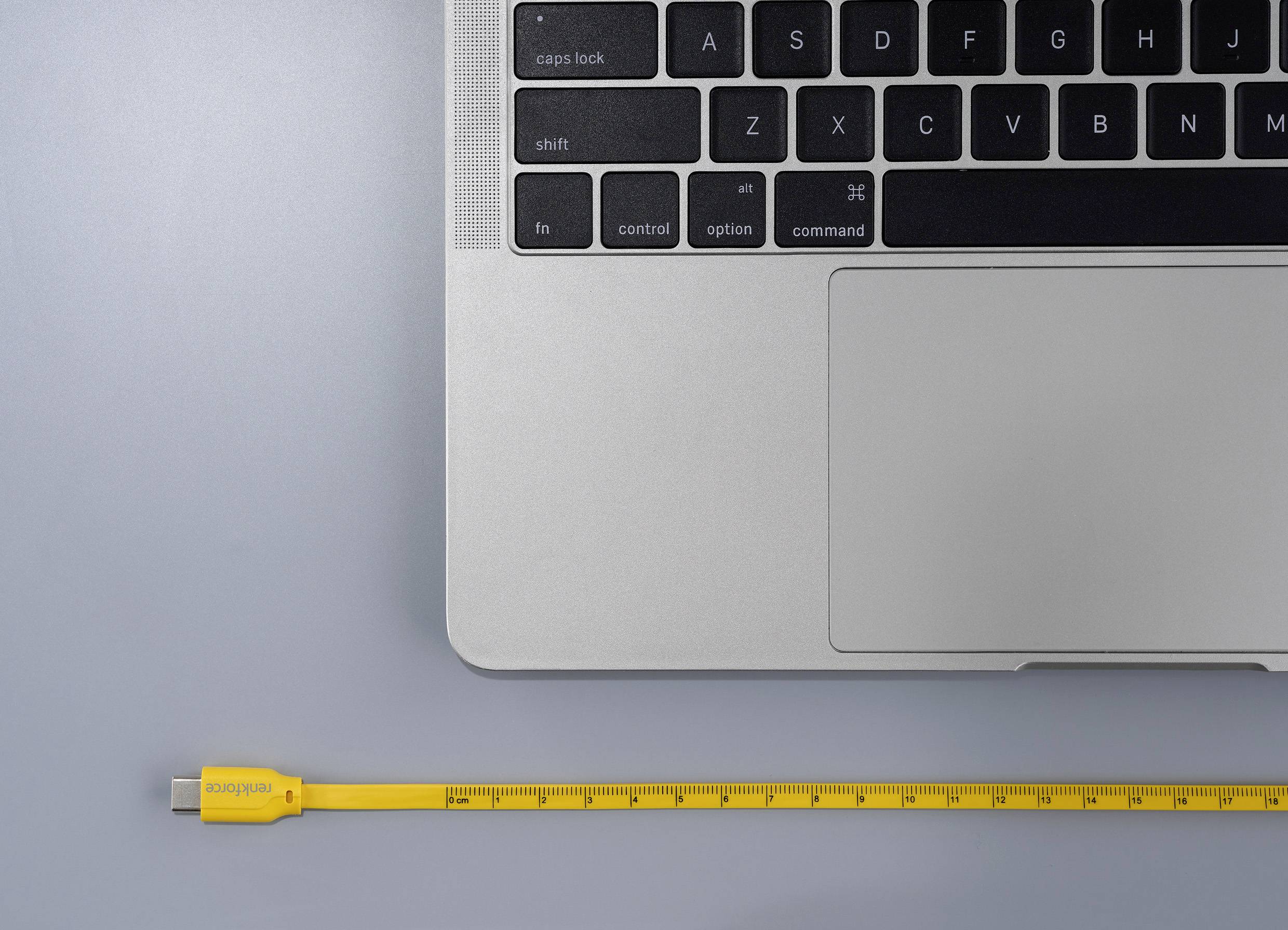 Ein Laptop mit Tastatur auf grauem Hintergrund. Daneben liegt ein gelbes USB-Kabel, das wie ein Maßband gestaltet ist.