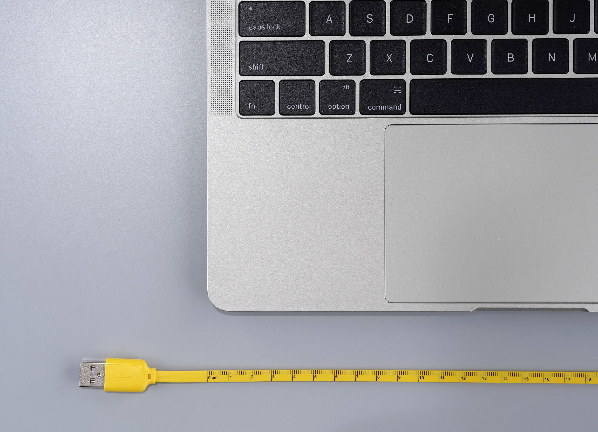 Laptop-Tastatur in der Ecke oben rechts, gelbes USB-Kabel mit Zentimeter-Markierungen darunter auf grauem Hintergrund.
