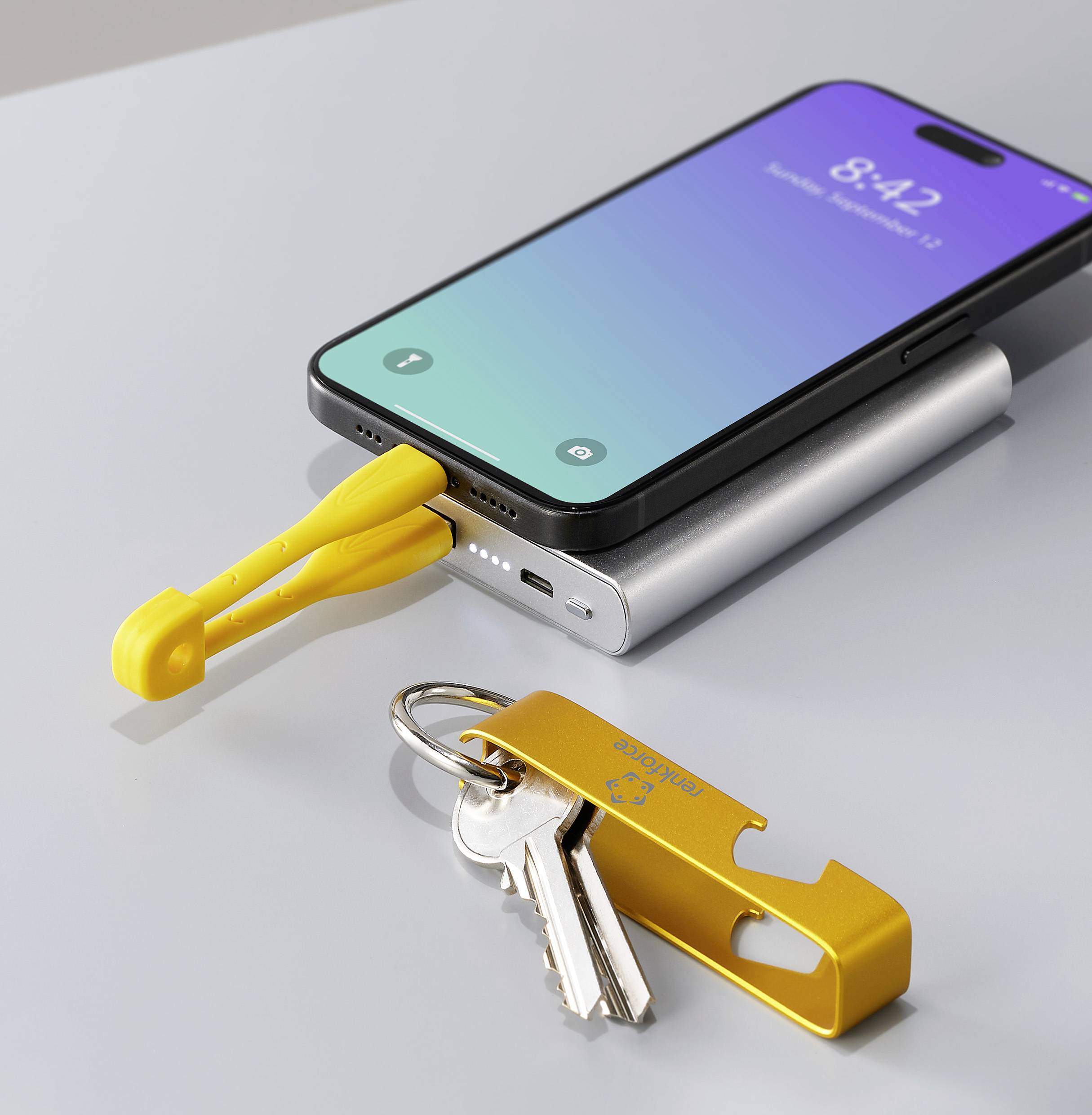 Ein Smartphone wird mit einem gelben USB-C-Kabel an eine Powerbank angeschlossen. Daneben liegt ein Schlüsselbund mit einem gelben Schlüsselanhänger.