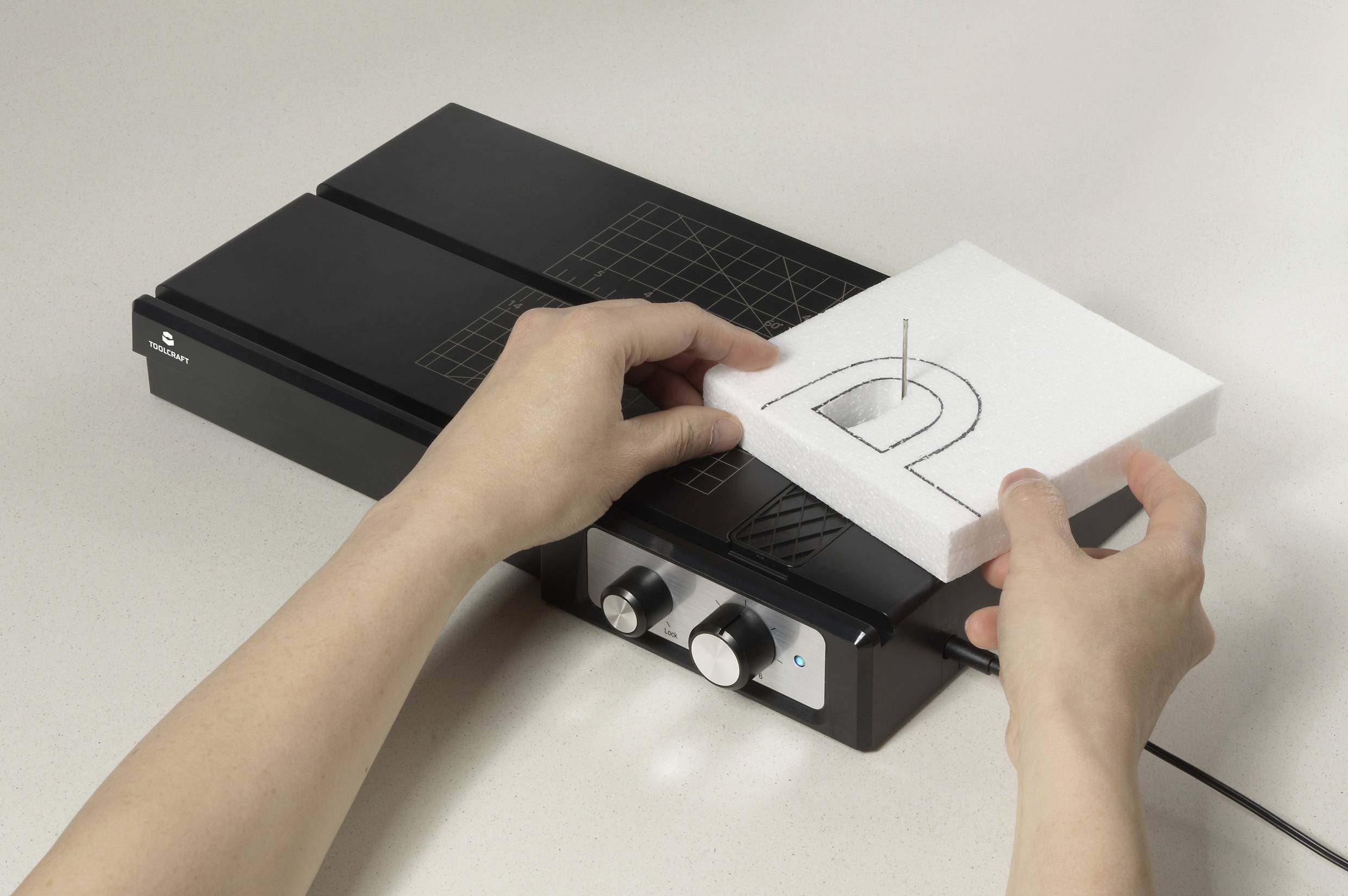 Zwei Hände schneiden mit einem Heißdrahtschneider einen Buchstaben 'D' aus weißem Schaumstoff.