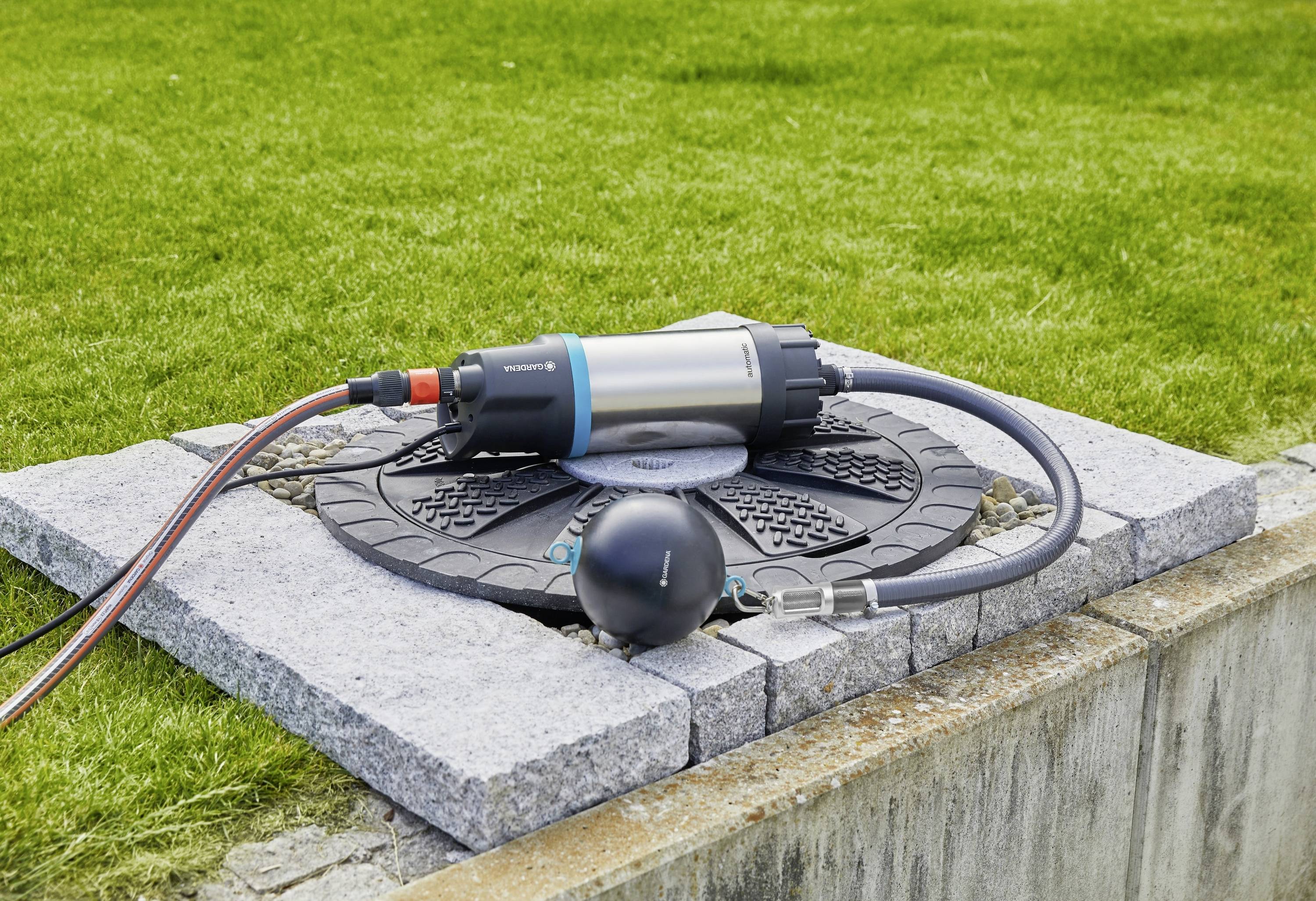Wasserpumpe auf einer Betonplatte in einem Garten; Schläuche sind angeschlossen, um Wasser zu fördern. Rasen im Hintergrund.