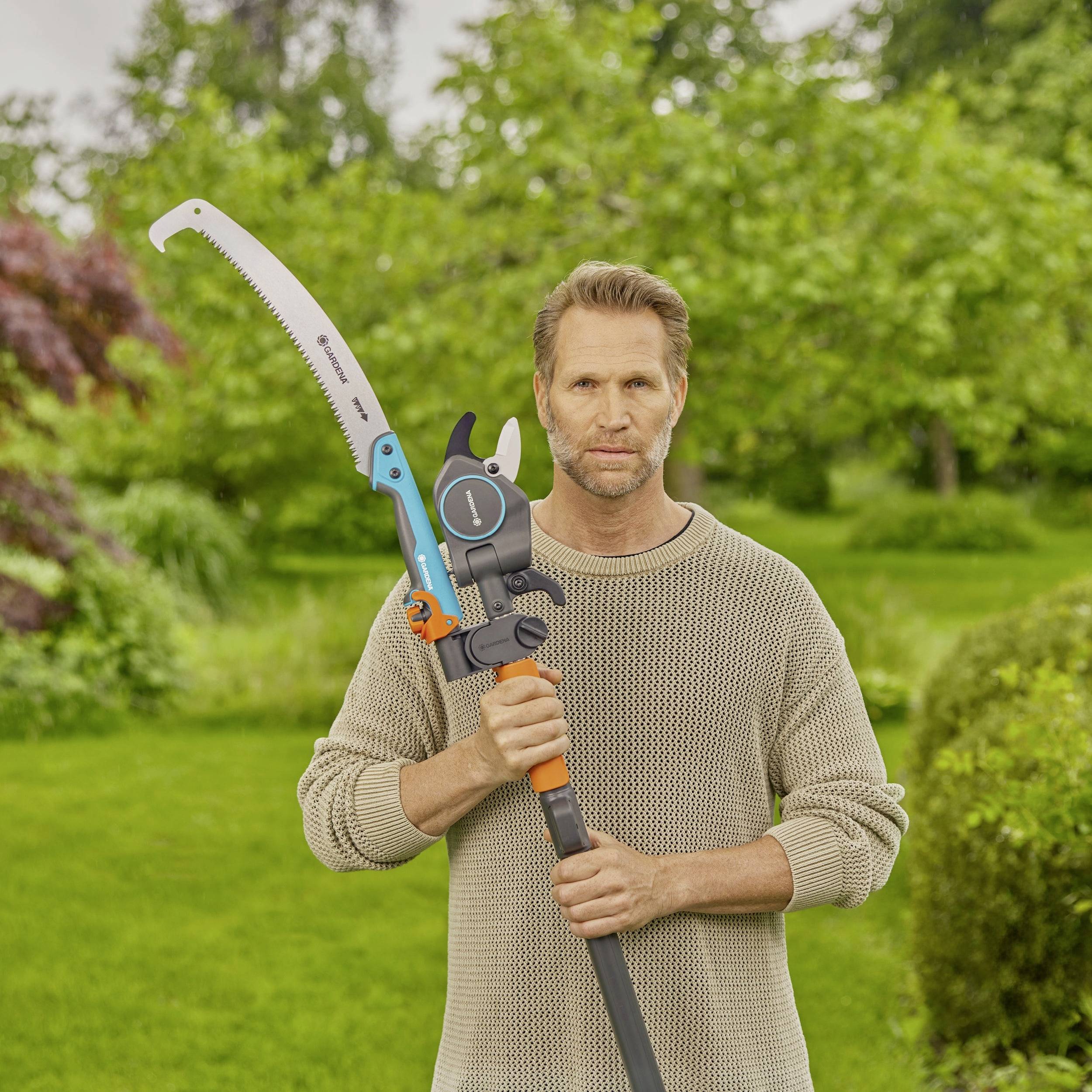 Ein Mann steht in einem Garten und hält eine Astsäge mit oranger und grauer Farbe. Im Hintergrund sind Bäume und Büsche zu sehen.