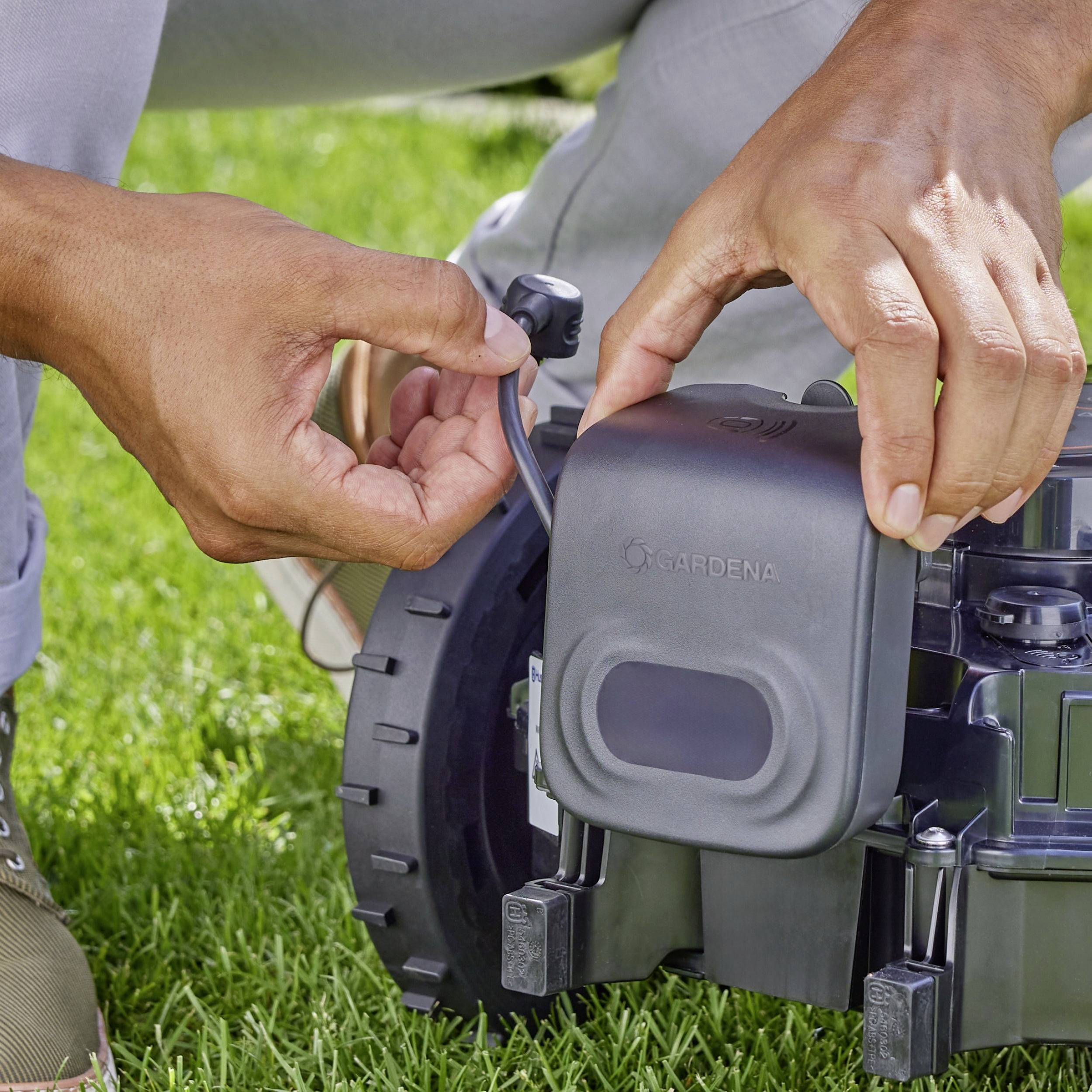Person installiert einen Mähroboter im Garten. Hände schließen ein Kabel an das Gerät auf dem Rasen an.
