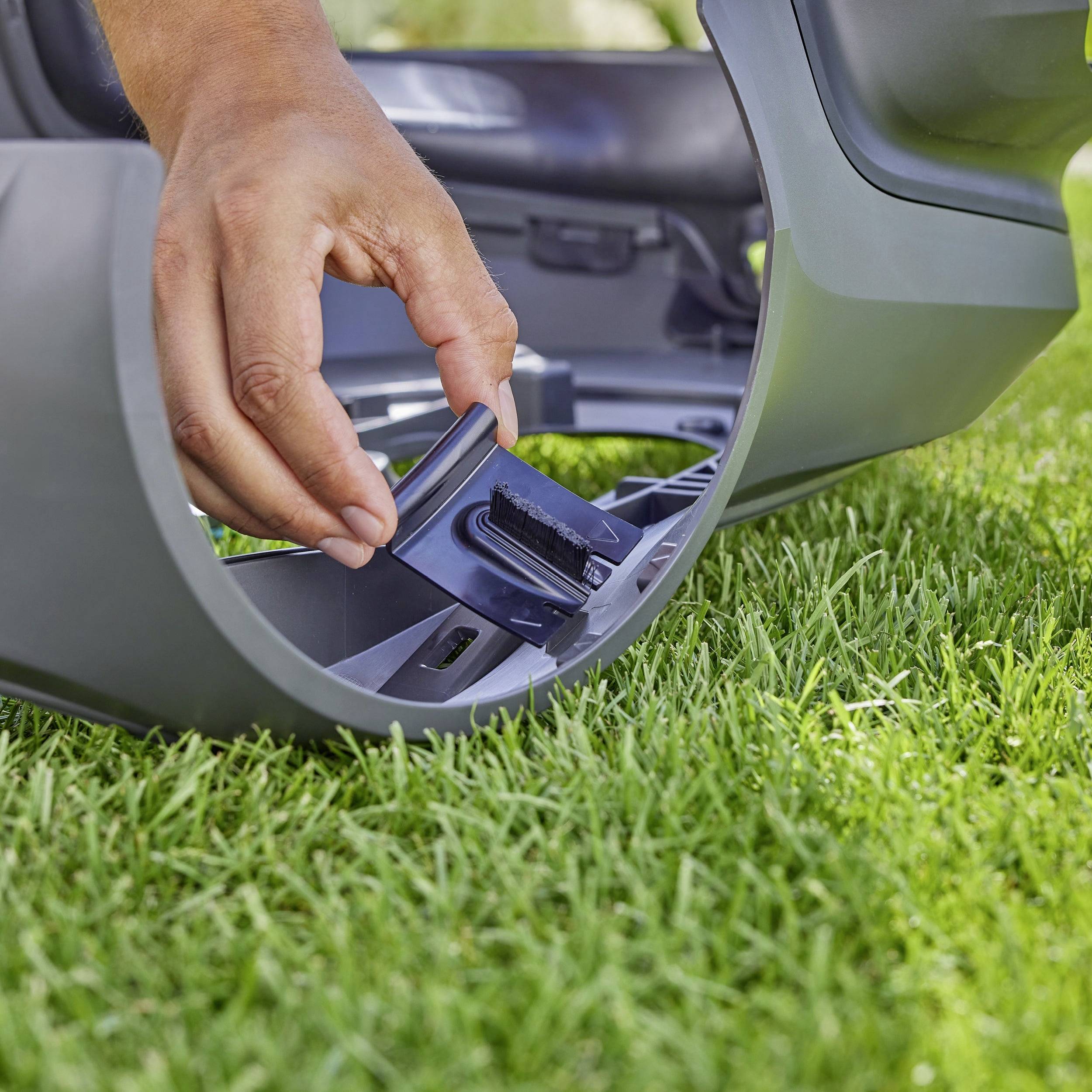 Eine Person hält ein Ersatzteil eines Rasenroboters auf einer grünen Wiese, zeigt das Einsetzen oder Entfernen eines Teils aus dem Gerät.