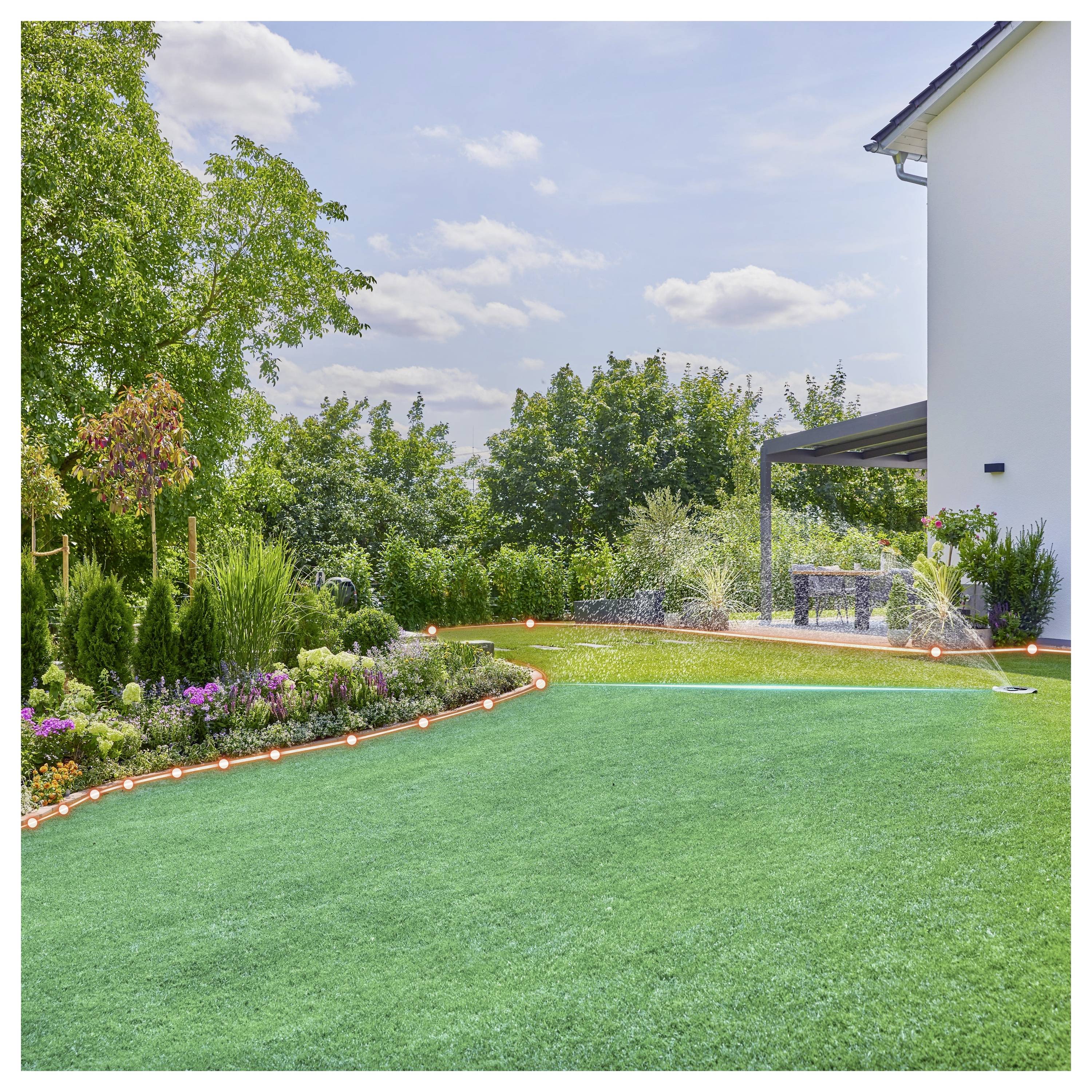 A well-maintained garden with green grass, a flower bed, and a hose watering the lawn adjacent to a modern house under a clear sky.