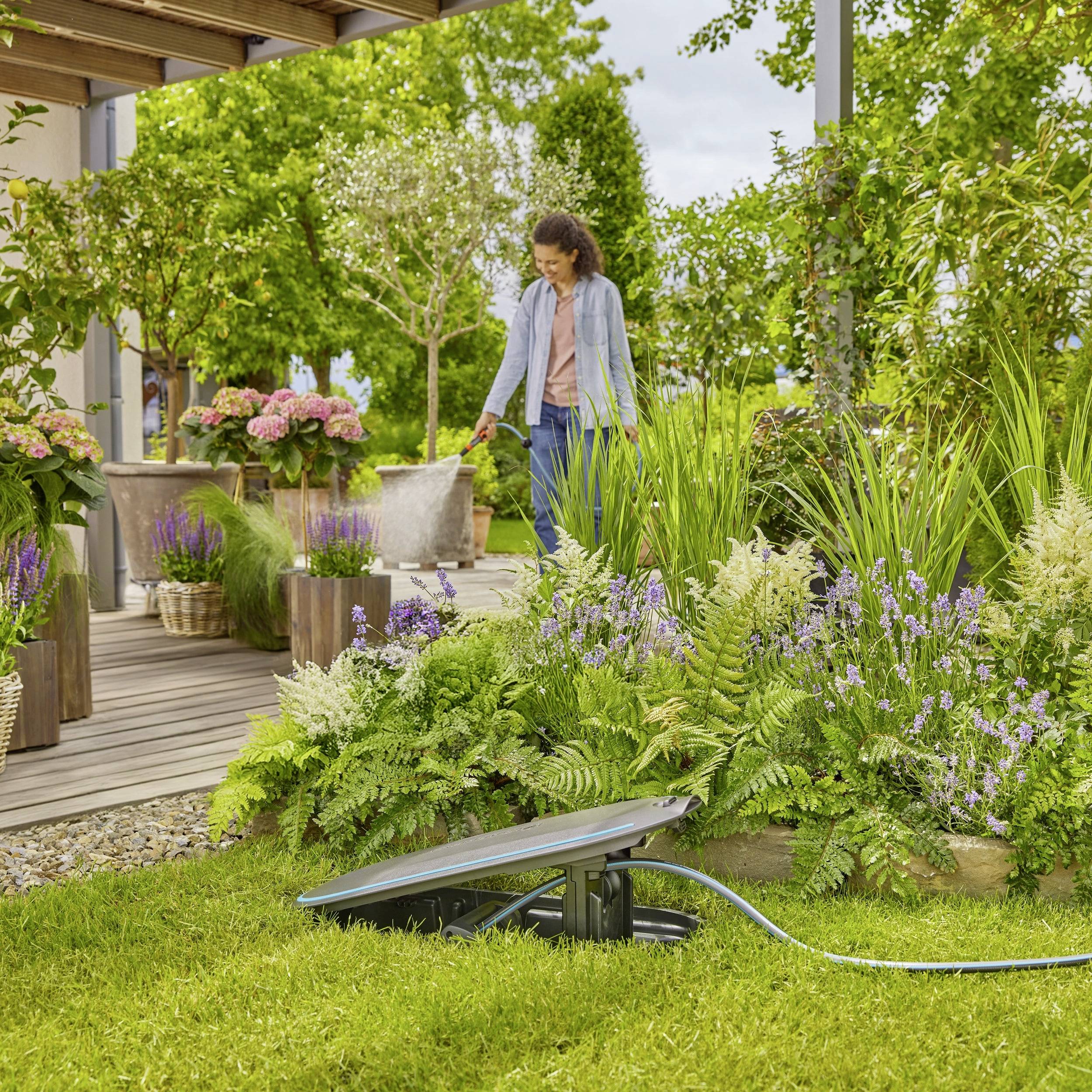 Eine Person steht in einem Garten neben Blumen und Pflanzen, umgeben von grünem Laub. Ein Solarstrahler ist im Vordergrund zu sehen.