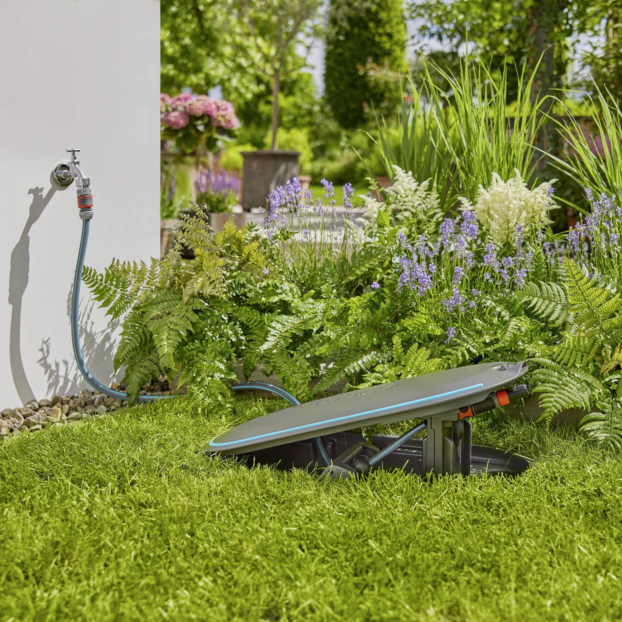 Rasen im Garten mit einem versenkbaren Rasenmäher nahe einer Wasserquelle, umgeben von Pflanzen und Blumen bei sonnigem Wetter.