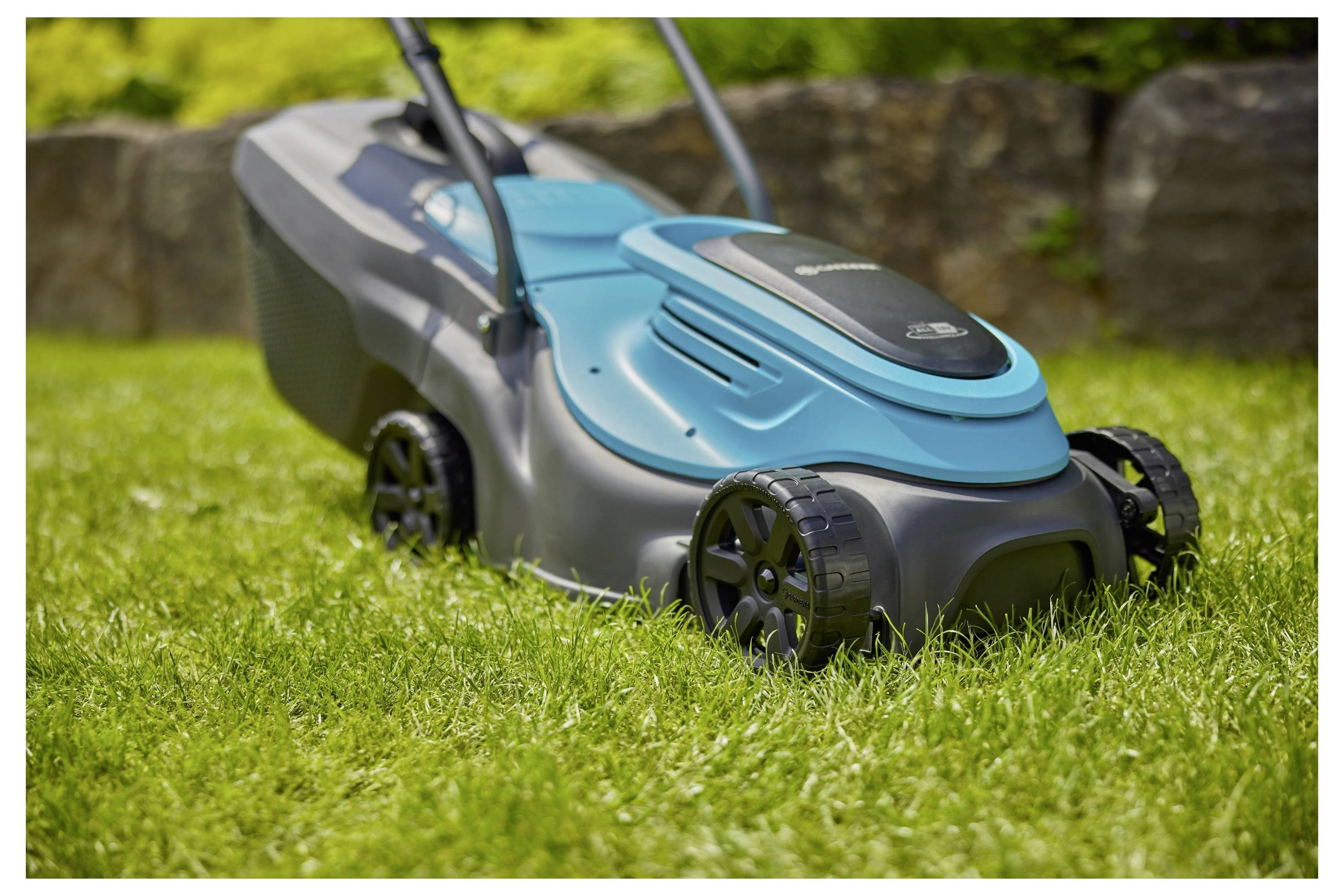 Eine Person mäht einen üppig grünen Rasen mit einem modernen, kleinen Rasenmäher. Dichte Büsche und Bäume sind im Hintergrund unter einem bewölkten Himmel.