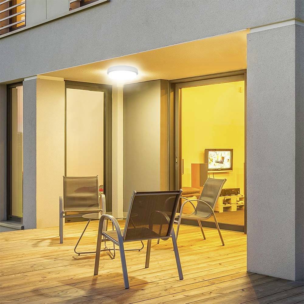 Balkon eines modernen Hauses bei Nacht, erhellt von einer Außenlampe; zwei Stühle und ein Tisch im Vordergrund, Fenster zeigt beleuchteten Innenraum.