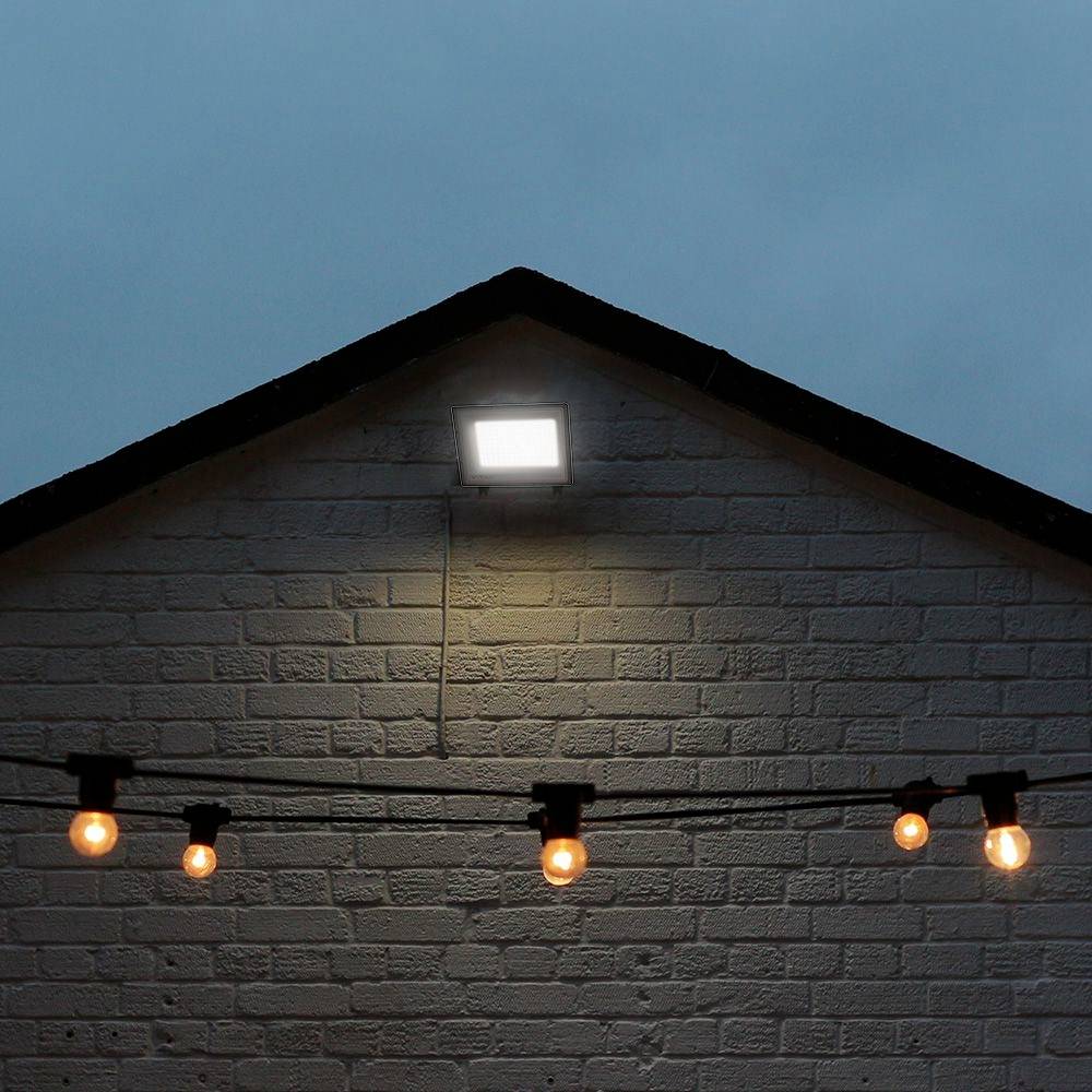 Ein weiß gestrichenes Giebelhaus mit einem Außenstrahler, der den oberen Teil beleuchtet. Im Vordergrund hängen Lichterketten mit gelben Glühbirnen.