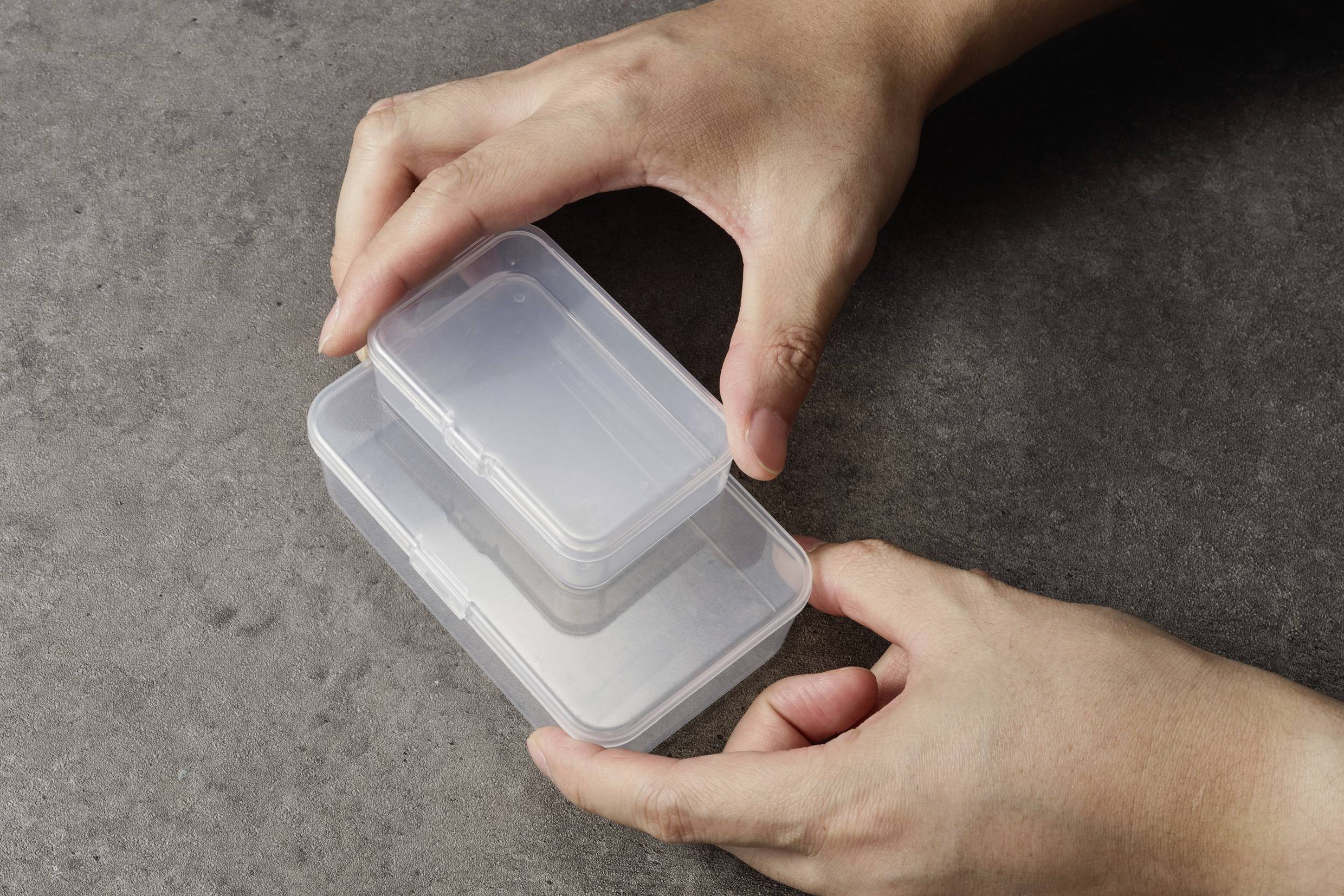 Zwei Hände stapeln transparente rechteckige Plastikbehälter auf einer grauen Oberfläche.