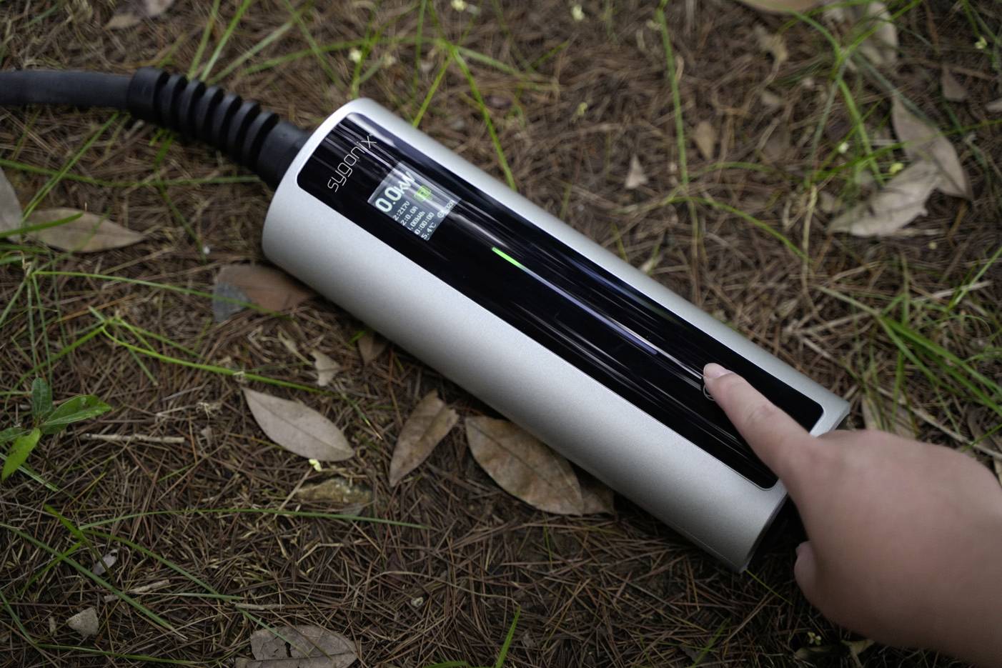 Ein Hand zeigt auf eine an einem Kabel befestigte Ladesäule, die auf einem Waldboden liegt. Nutzung für Elektrofahrzeuge angezeigt.