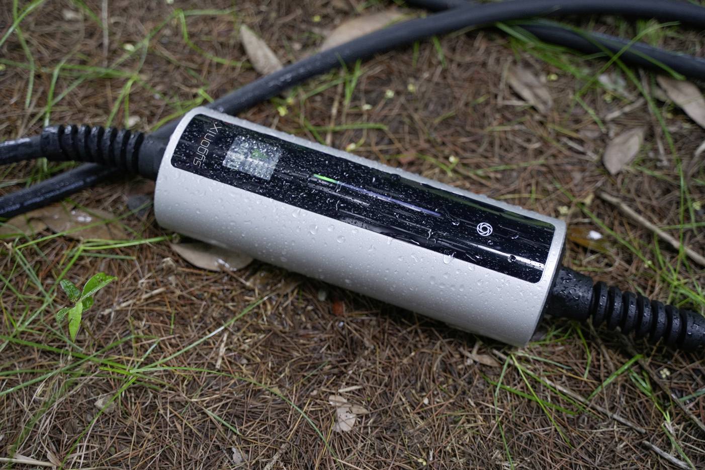 Ein elektrisches Ladekabel liegt auf dem Boden im Freien, gezeigt mit einem AC-Ladegerät, das an ein schwarzes Kabel angeschlossen ist.