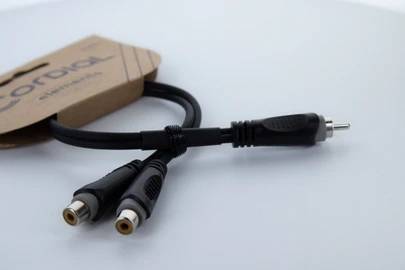 Schwarz-Weiß-Cinch-Y-Kabel mit zwei Buchsen und einem Stecker, auf einer weißen Oberfläche liegend, verbunden mit Pappverpackung.