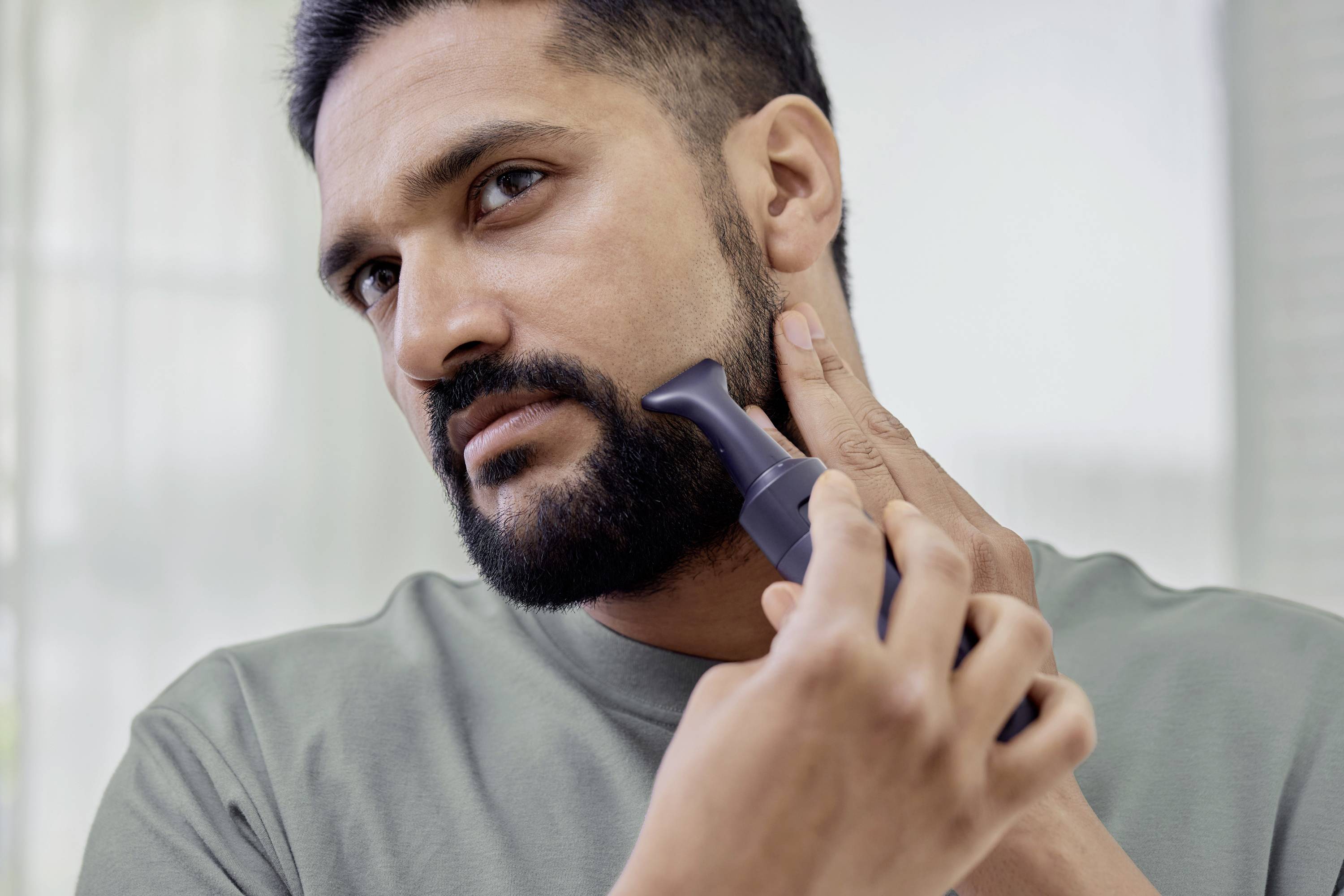 Ein Mann nutzt einen elektrischen Trimmer, um seinen Bart zu pflegen, in einem hell beleuchteten Raum.