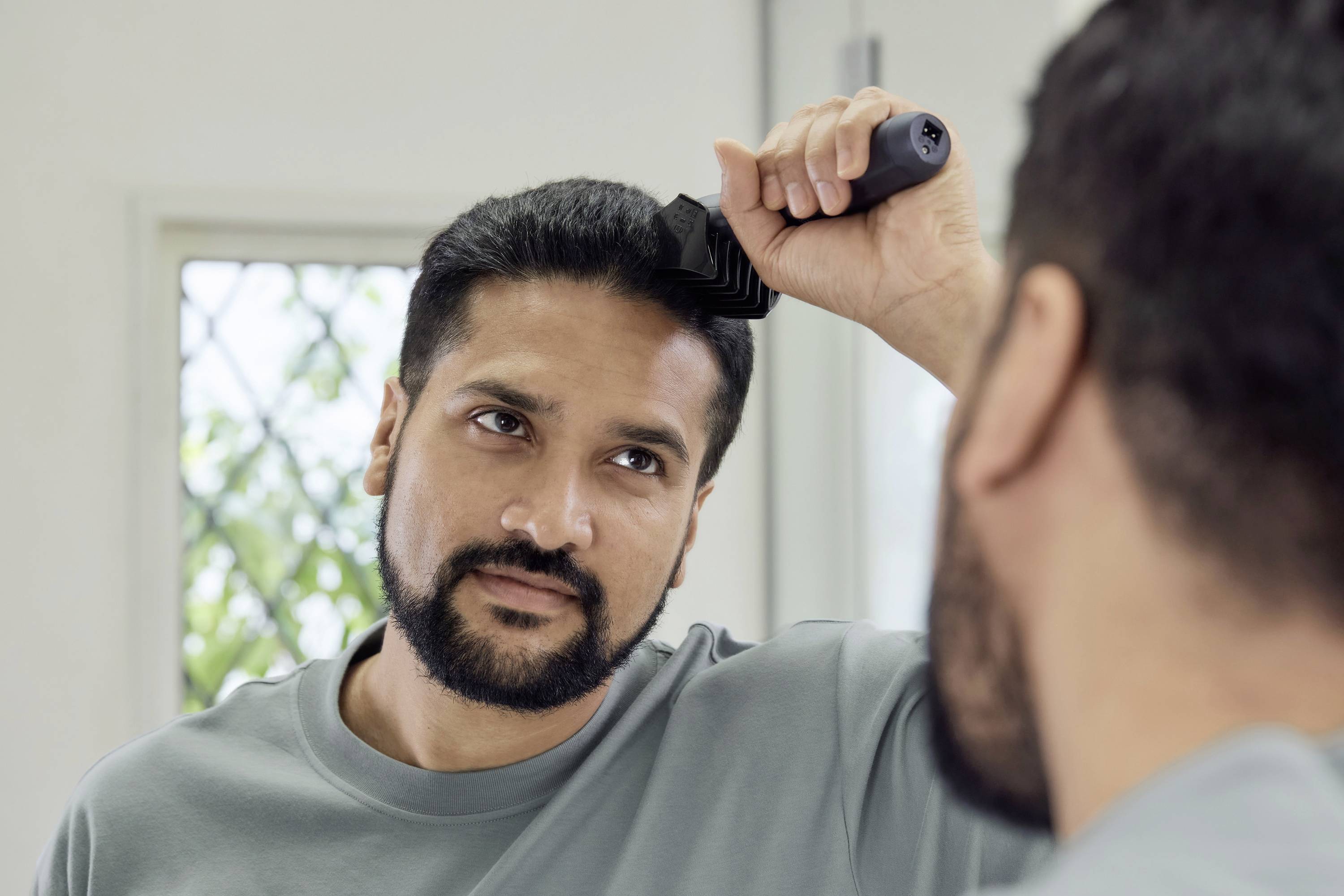 Ein Mann kämmt sein Haar vor einem Spiegel. Er trägt ein graues Shirt. Im Hintergrund ist ein Fenster mit einem leicht verschwommenen Blick nach draußen.
