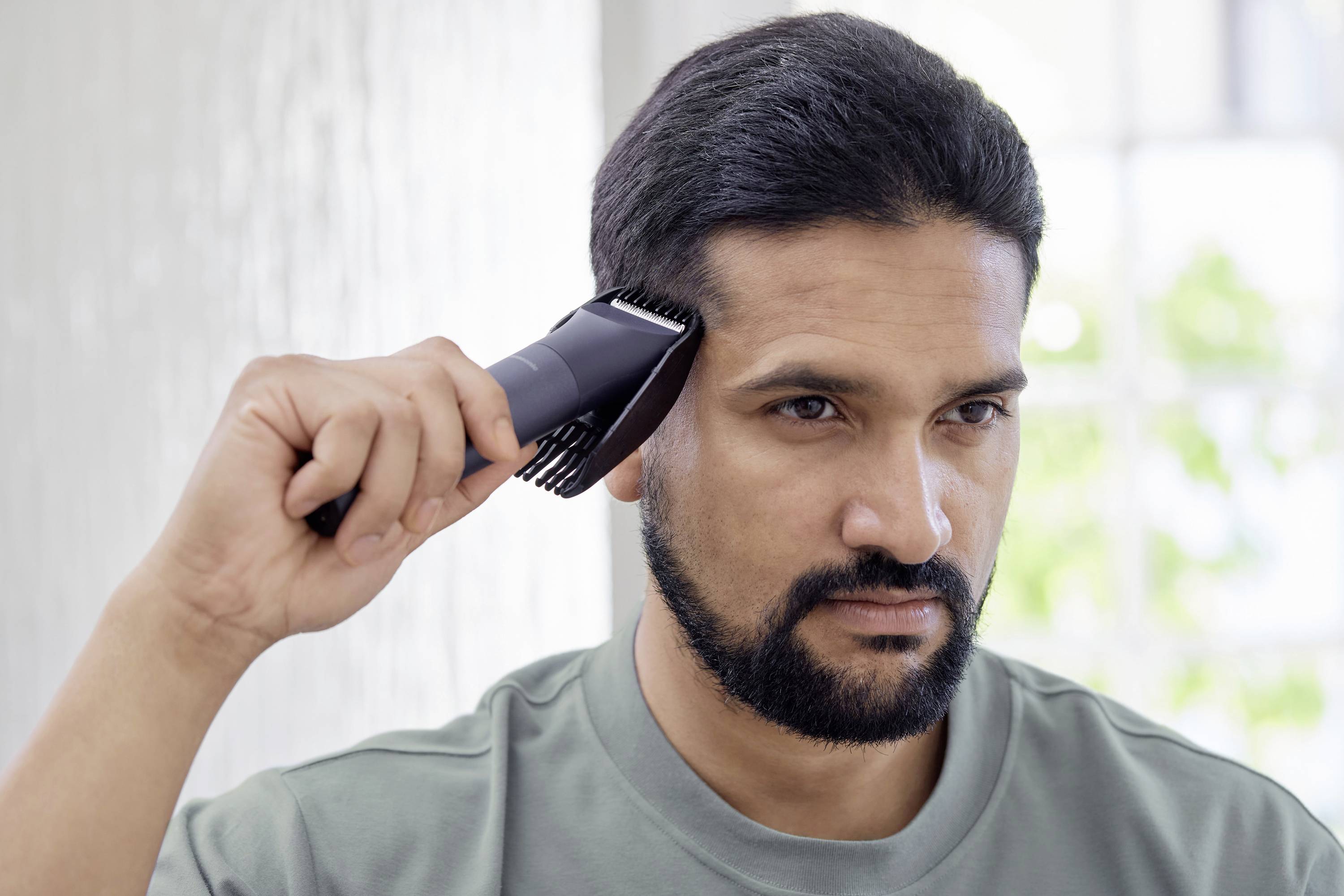 Ein Mann mit Bart kämmt sich die Haare mit einem elektrischen Haarschneider vor einem hellen Hintergrund.