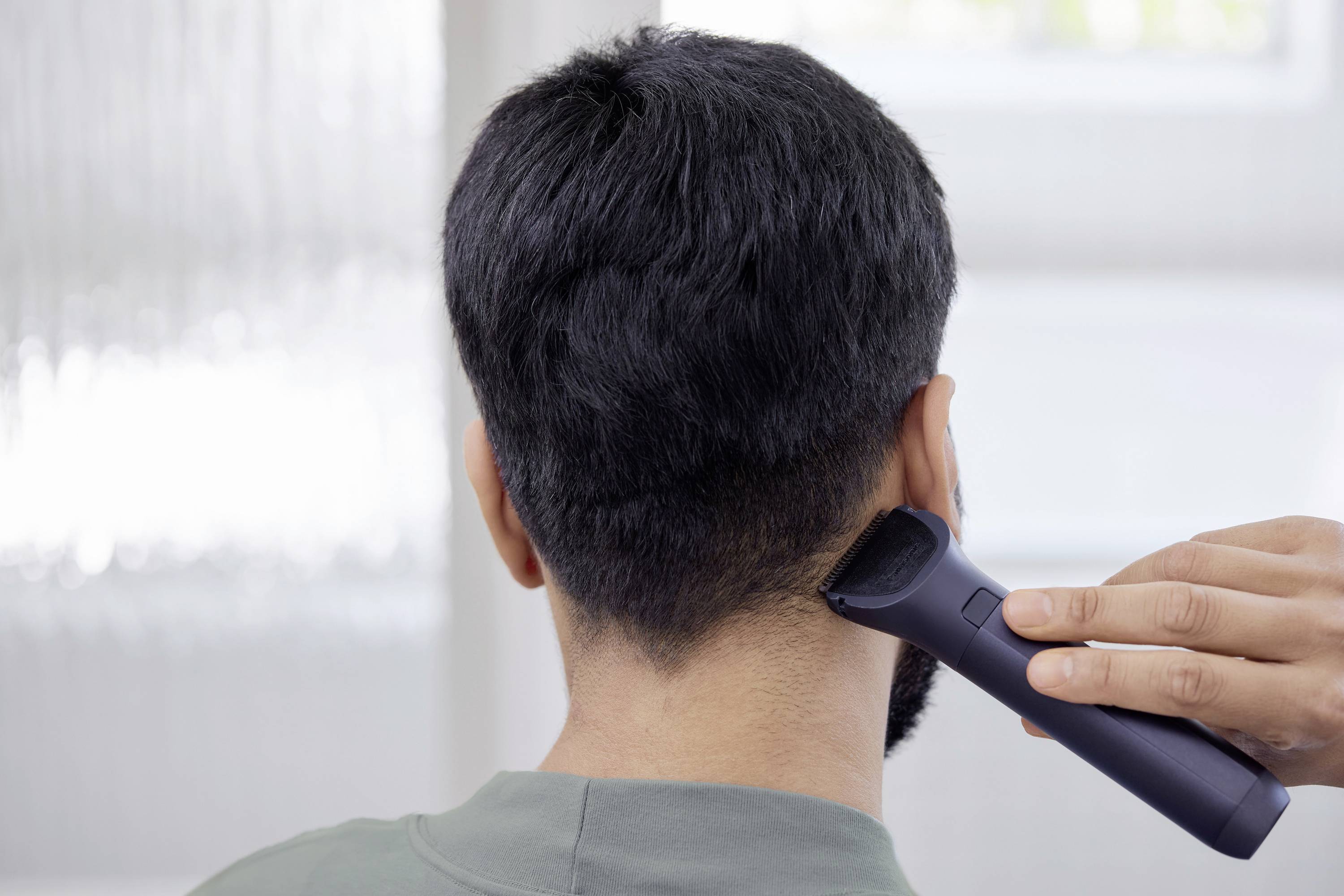 Person wird von hinten gezeigt, während die Haare mit einem elektrischen Rasierer getrimmt werden. Konzentration auf Nackenbereich.