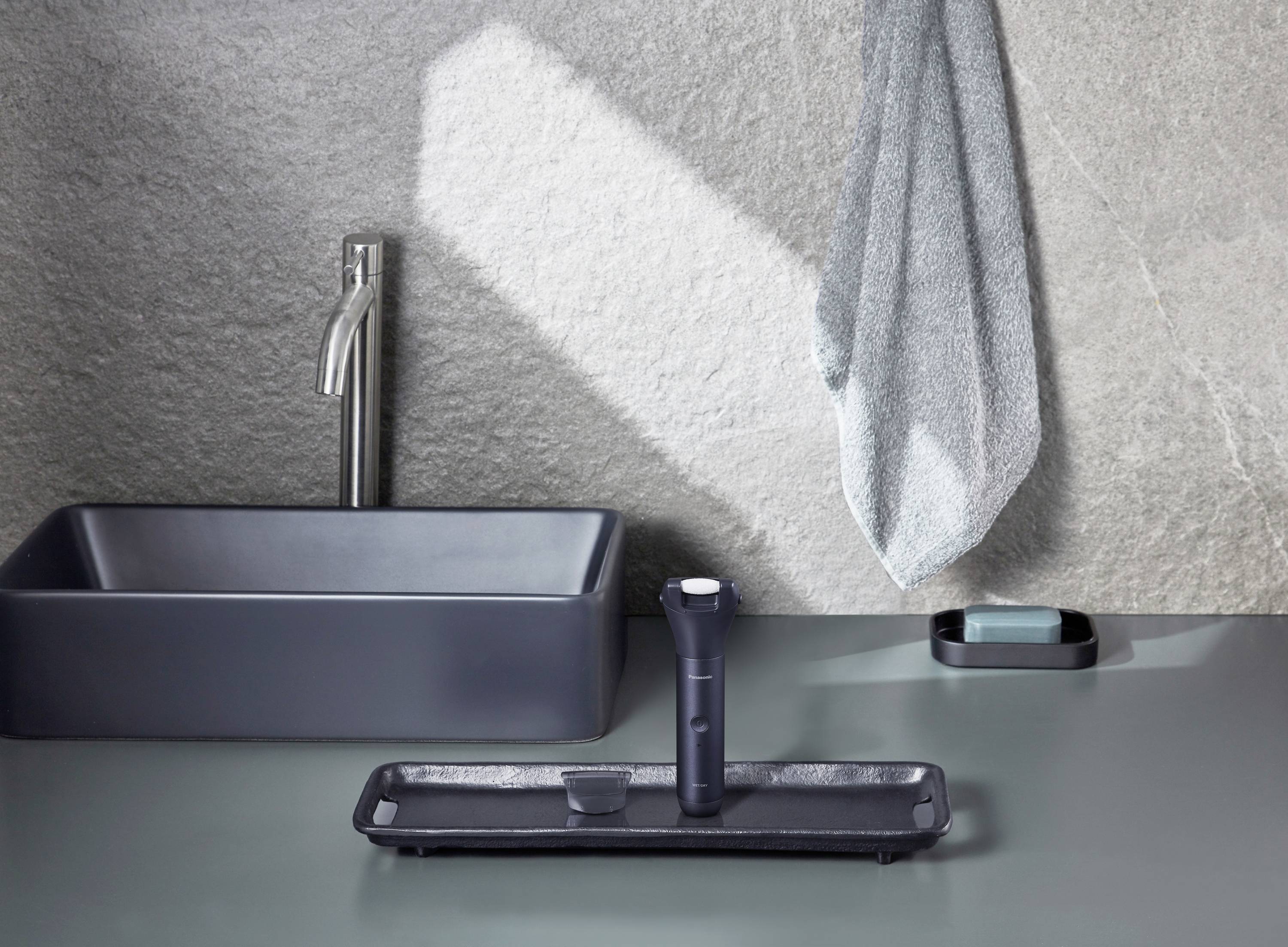 Ein graues Badezimmer mit einem Waschbecken links und einem Handtuch an der Wand. Auf dem Tisch stehen ein Rasierer und eine Seifenschale.