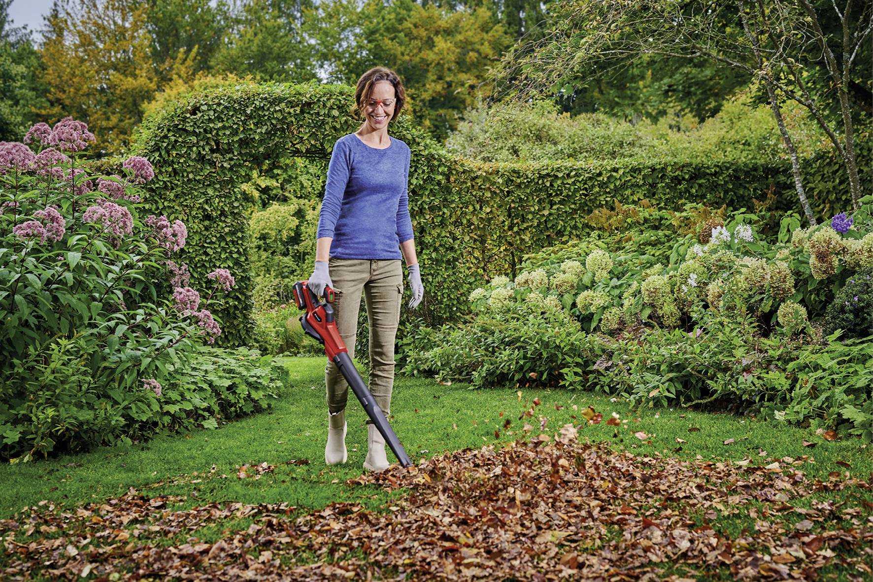 Eine Frau in Gartenkleidung benutzt einen Laubbläser, um einen Haufen Herbstlaub in einem üppigen Gartenbereich zu sammeln.