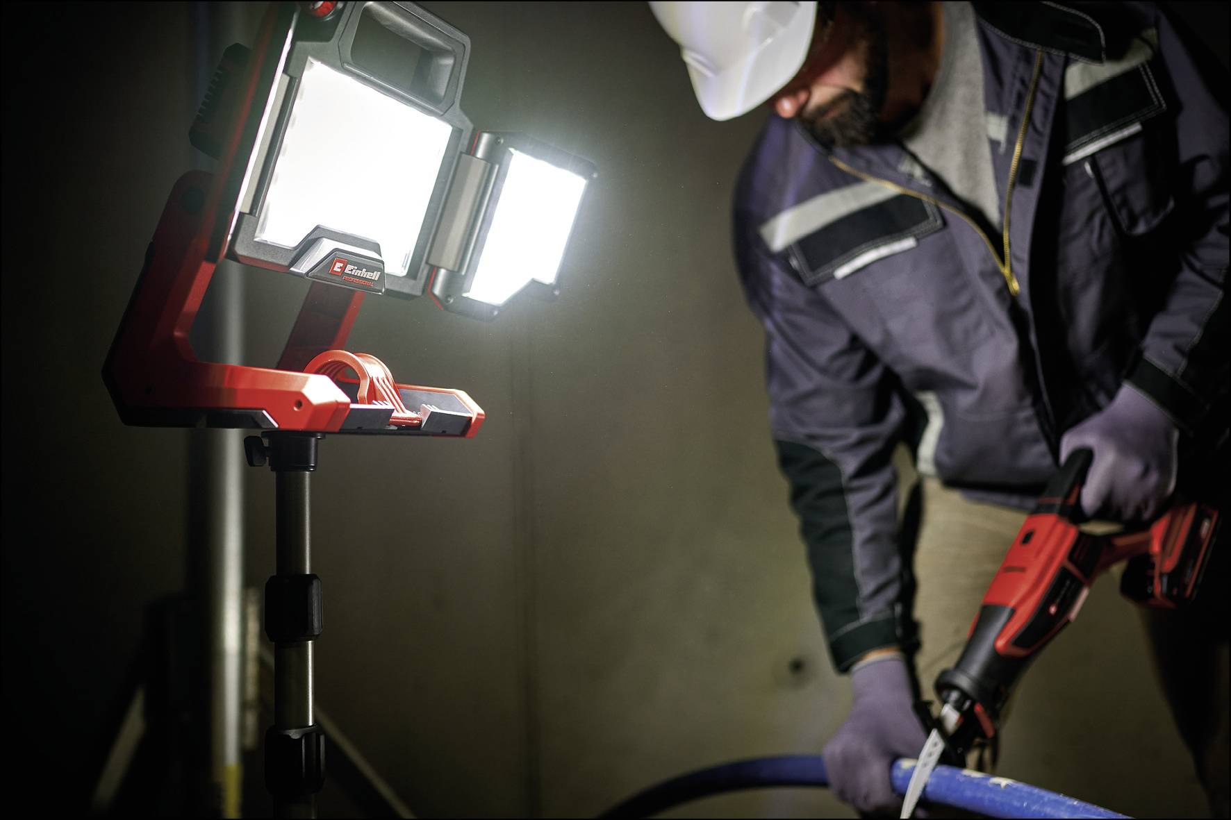 Ein Arbeiter mit Helm und Handschuhen verwendet ein Elektrowerkzeug. Eine hell leuchtende LED-Arbeitslampe beleuchtet die Szene.