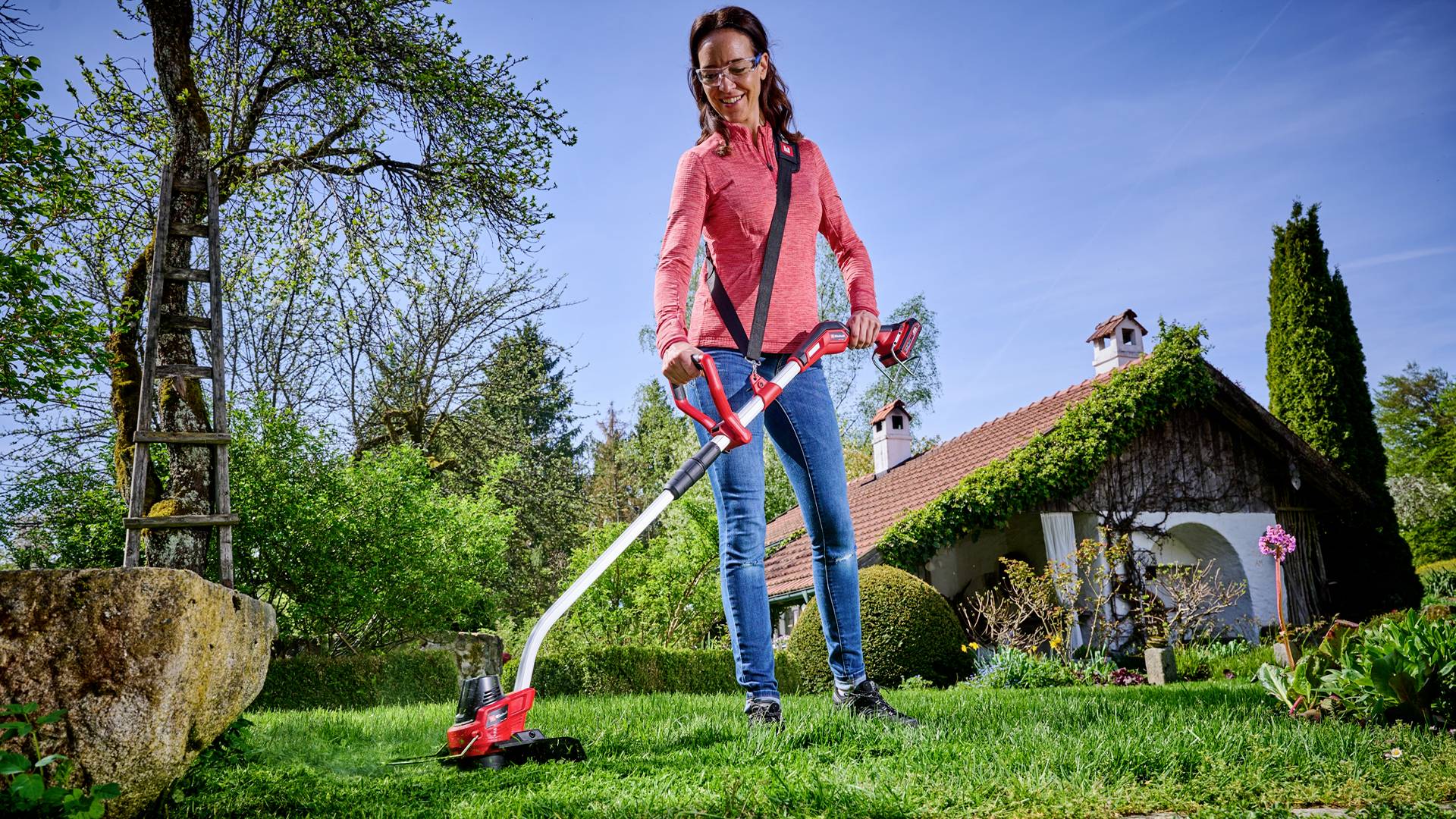 Eine Frau mäht mit einem roten Rasentrimmer den Rasen in einem Garten vor einem Haus mit rotem Dach. Im Hintergrund stehen Bäume.