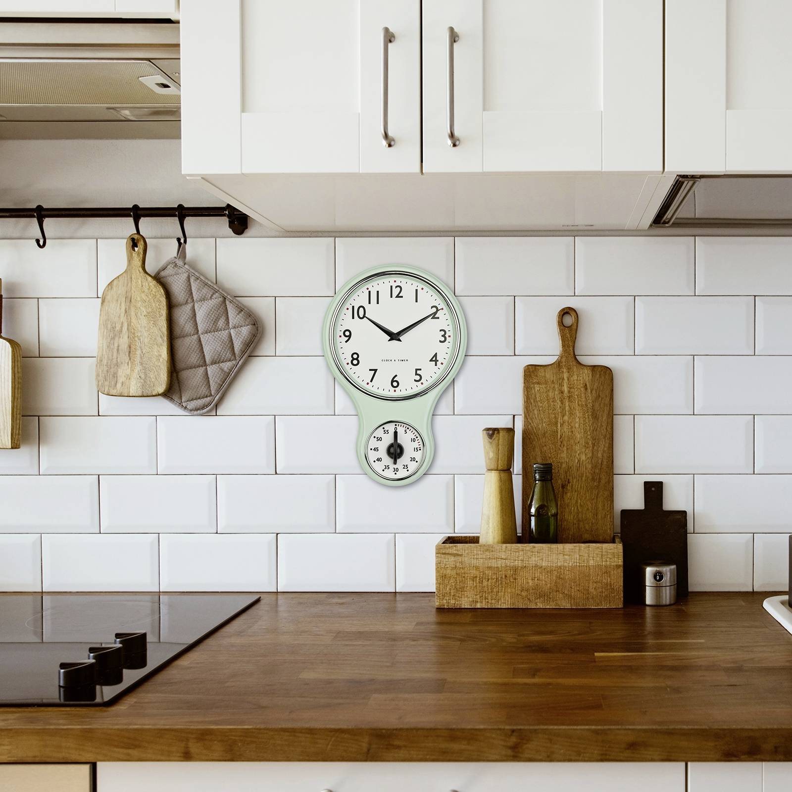 Eine Küche mit weiß gefliesten Wänden, hölzerner Arbeitsplatte, Schneidebrettern, Gewürzen und einer Wanduhr, die 10:09 Uhr zeigt.