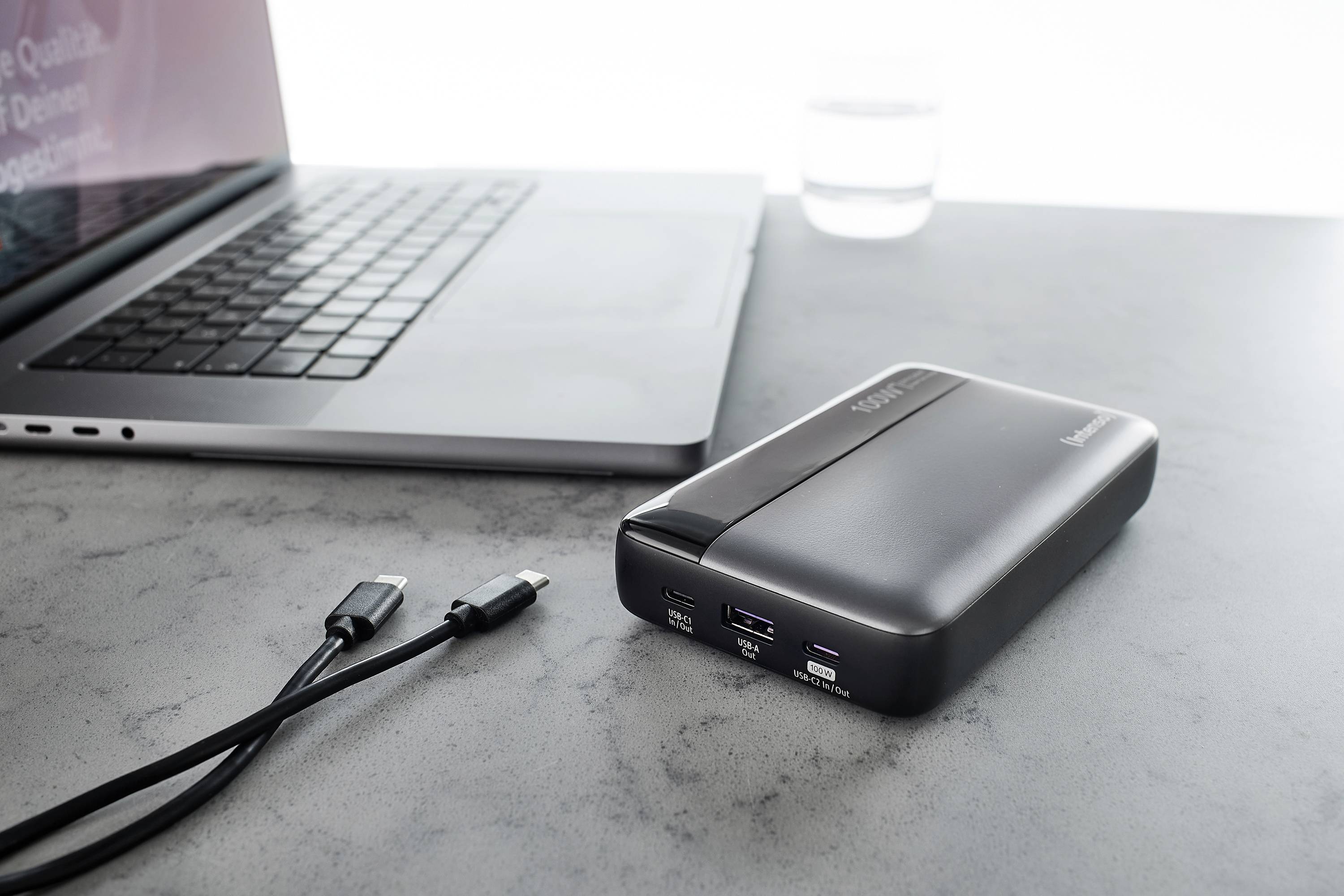 Eine Powerbank neben einem Laptop auf einem grauen Tisch, mit zwei USB-Kabeln daneben. Ideal für mobiles Laden von Geräten.