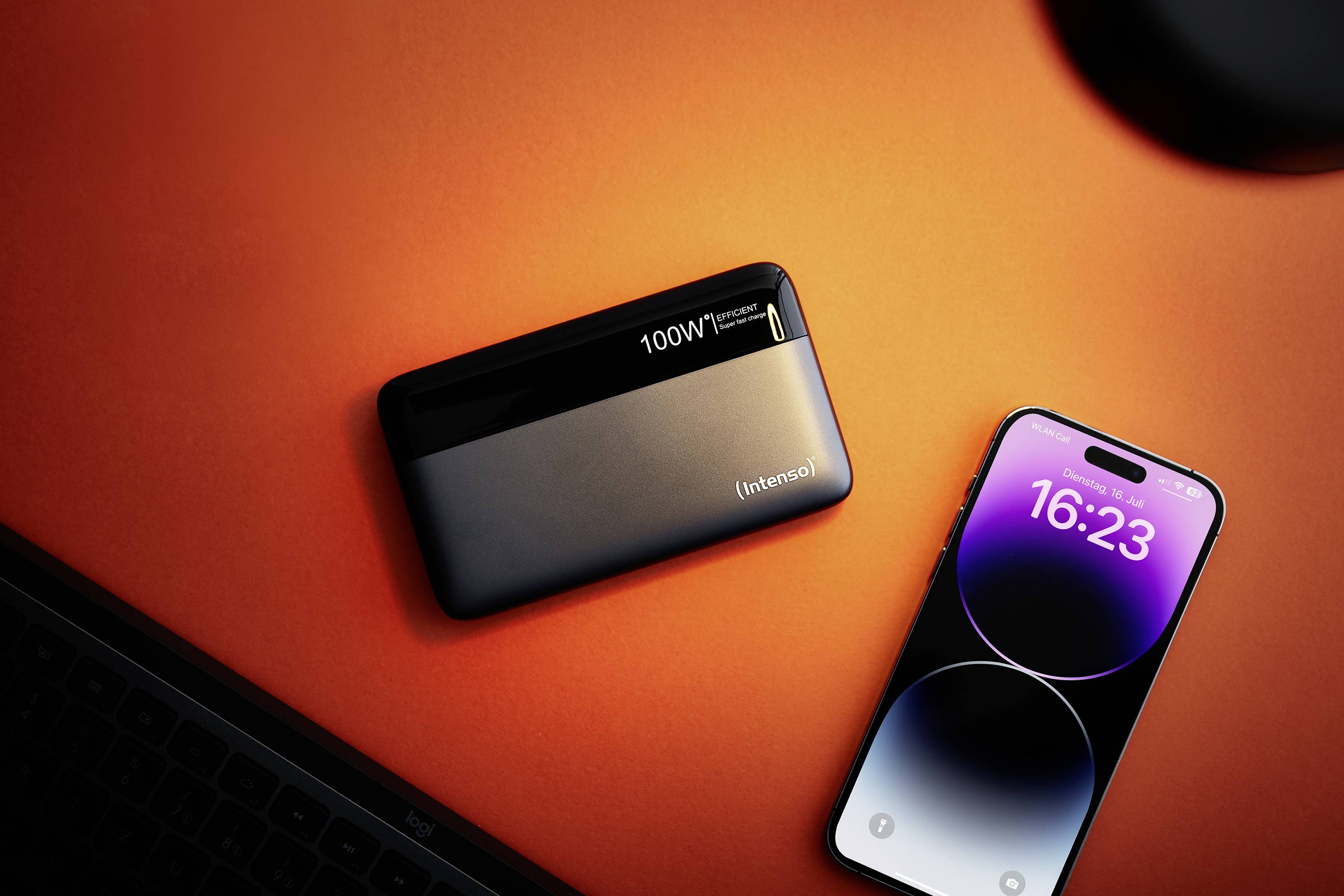 Ein Smartphone liegt neben einer schwarzen Powerbank auf einem orangefarbenen Tisch. Die digitale Uhrzeit auf dem Smartphone zeigt 16:23 an.