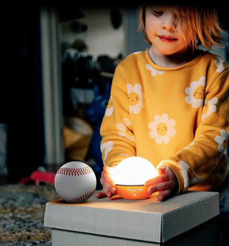 Ein Kind in einem gelben Pulli mit Blumenmuster hält ein leuchtendes Nachtlicht auf einem Karton, daneben liegt ein Baseball.