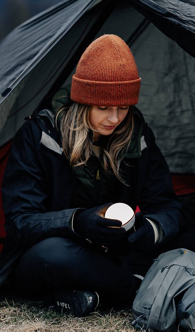 Eine Person sitzt vor einem Zelt und hält eine Tasse, gekleidet in warmer Outdoor-Kleidung samt Mütze und Handschuhen.