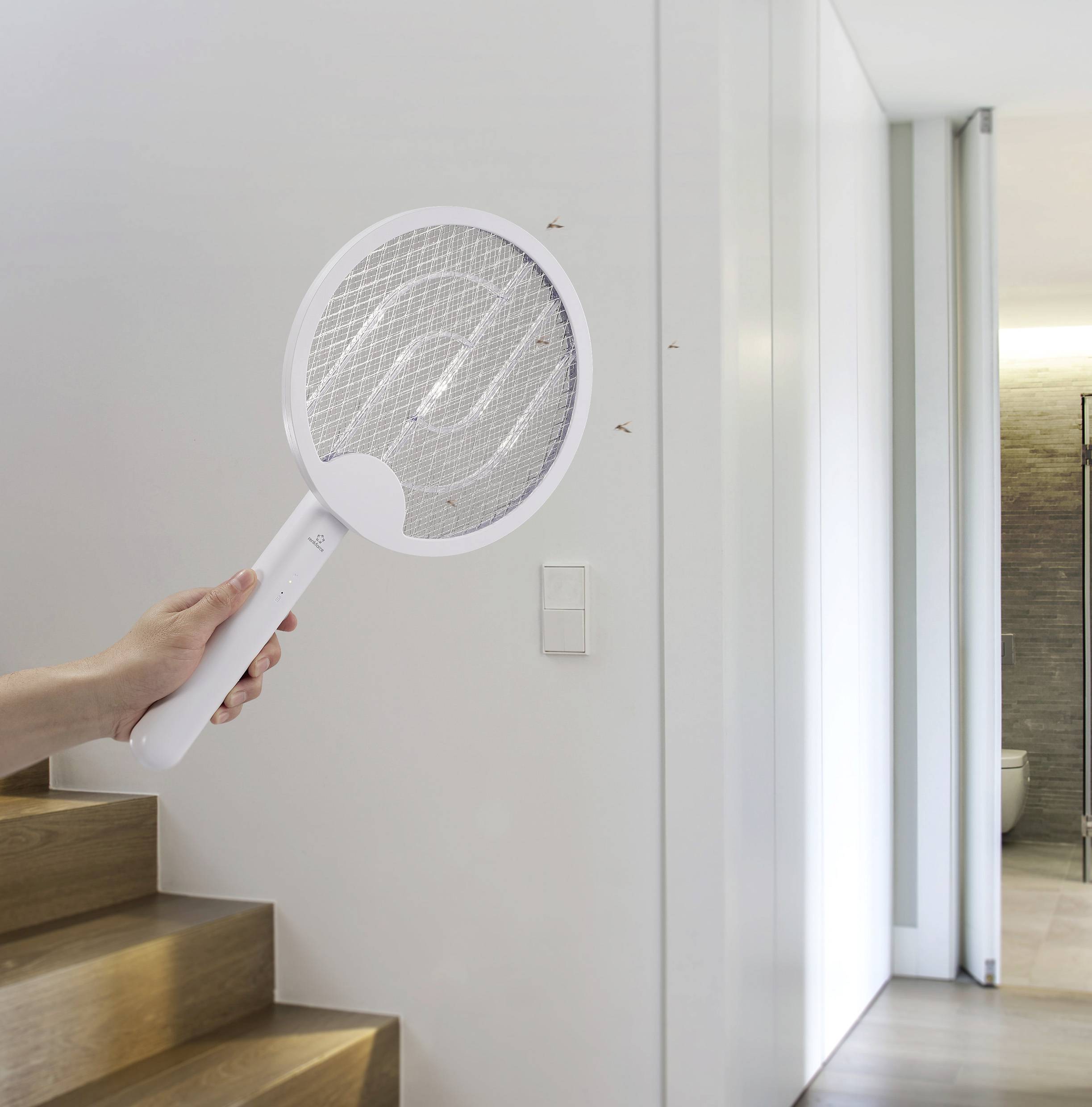 Eine Person hält eine elektrische Fliegenklatsche im Flur, um drei Fliegen an der Wand abzuwehren.