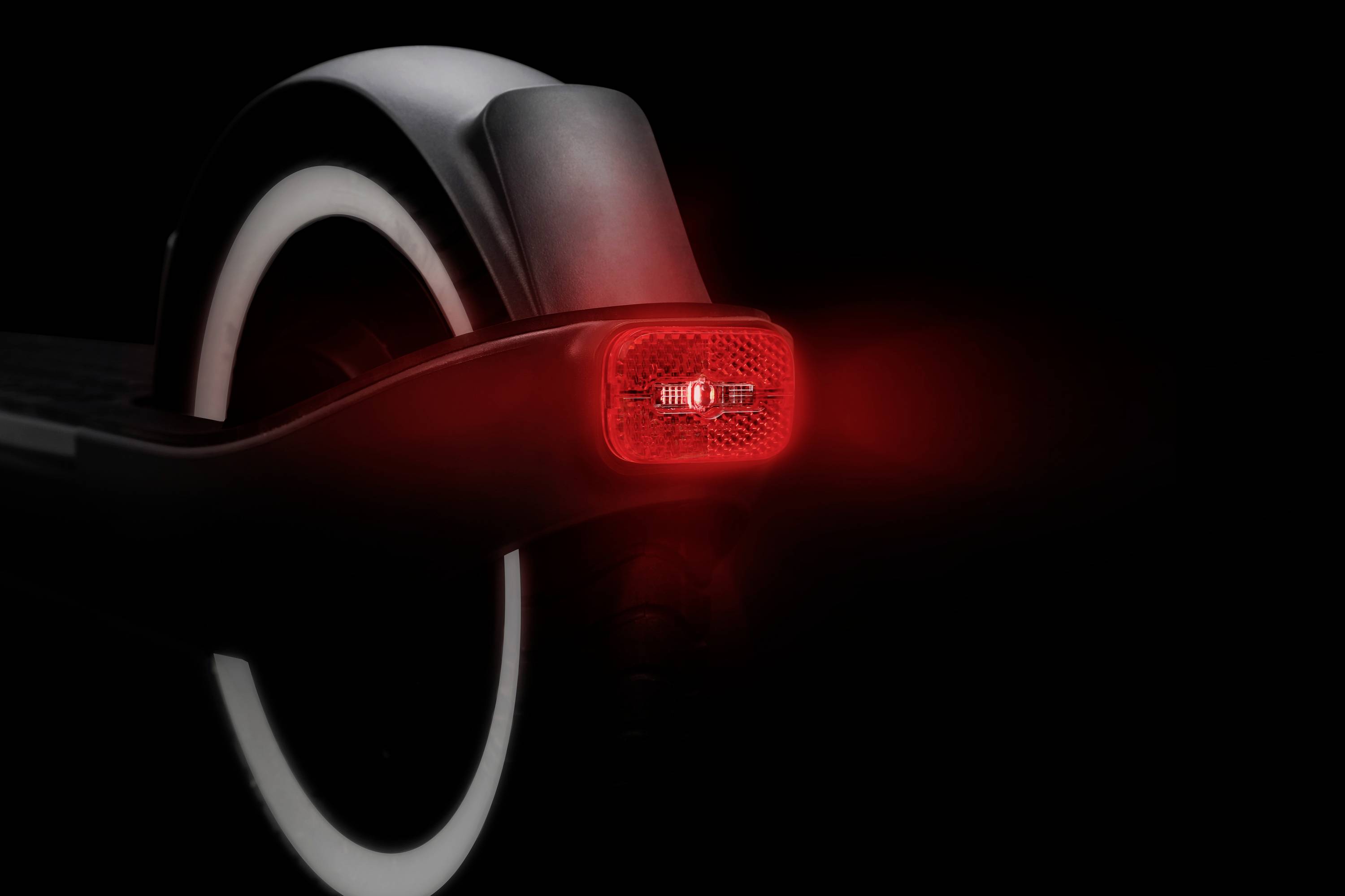 Ein elektrischer Roller von hinten, mit leuchtendem roten Rücklicht und reflektierender Oberfläche, auf schwarzem Hintergrund.