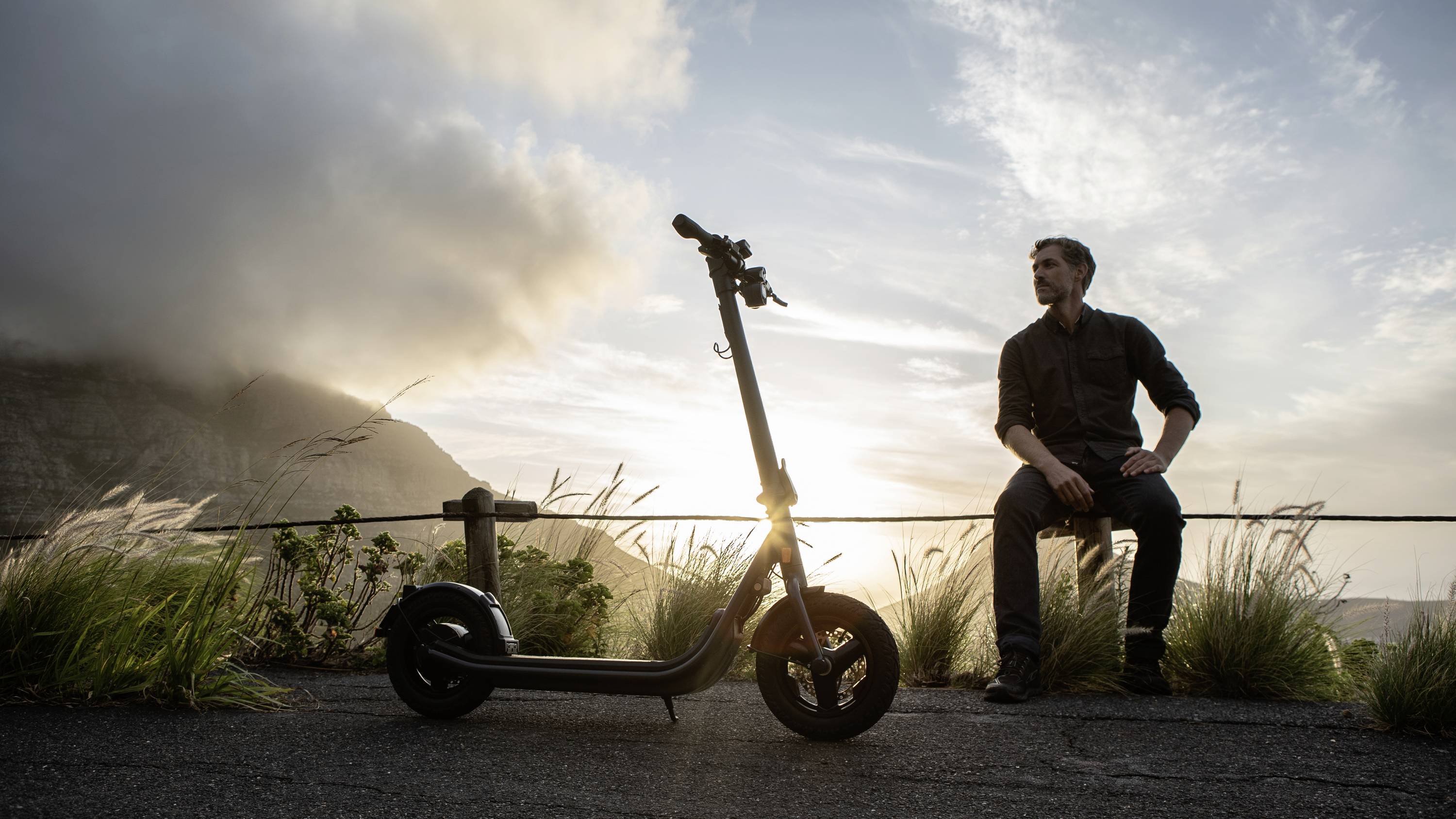 Ein Mann sitzt auf einer Mauer neben einem E-Roller bei Sonnenuntergang mit Bergen im Hintergrund, wirkt nachdenklich und ruhig.