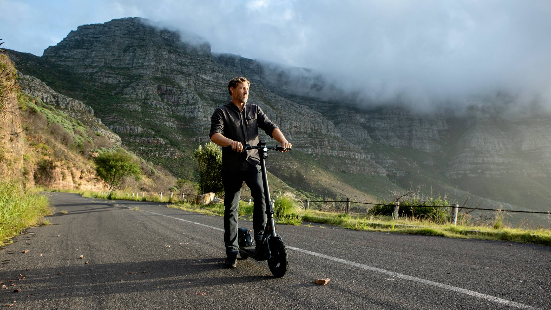 Ein Mann fährt auf einem elektrischen Roller eine leere Straße entlang, im Hintergrund sind bewaldete Berge und Wolken zu sehen.