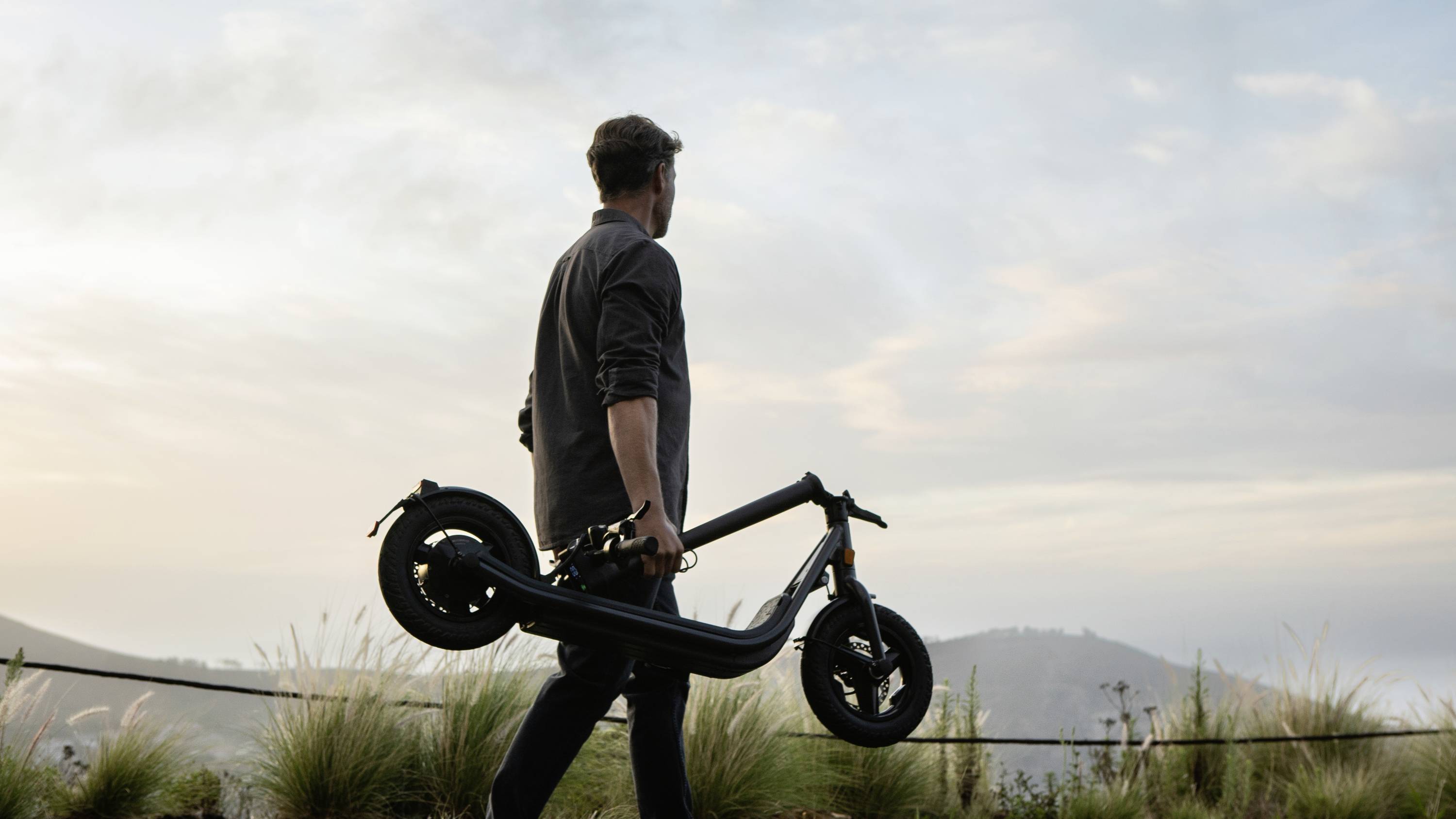Eine Person trägt einen Elektroroller und blickt in die Ferne, vor einem Panorama aus Hügeln und Himmel in der Abenddämmerung.