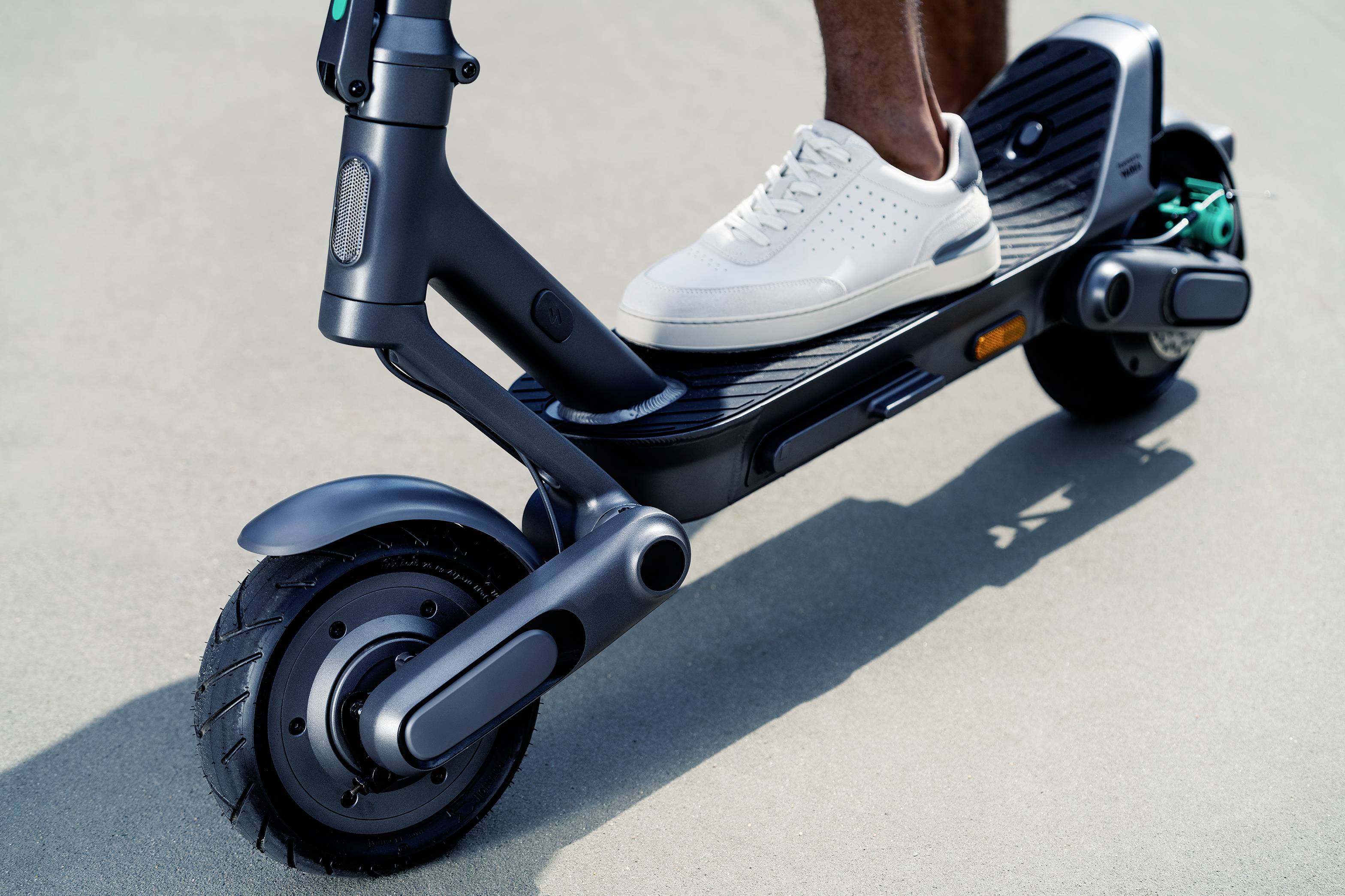 Eine Person steht mit einem Fuß auf einem Elektroroller. Der Roller hat schwarze Räder und ein modernes Design.