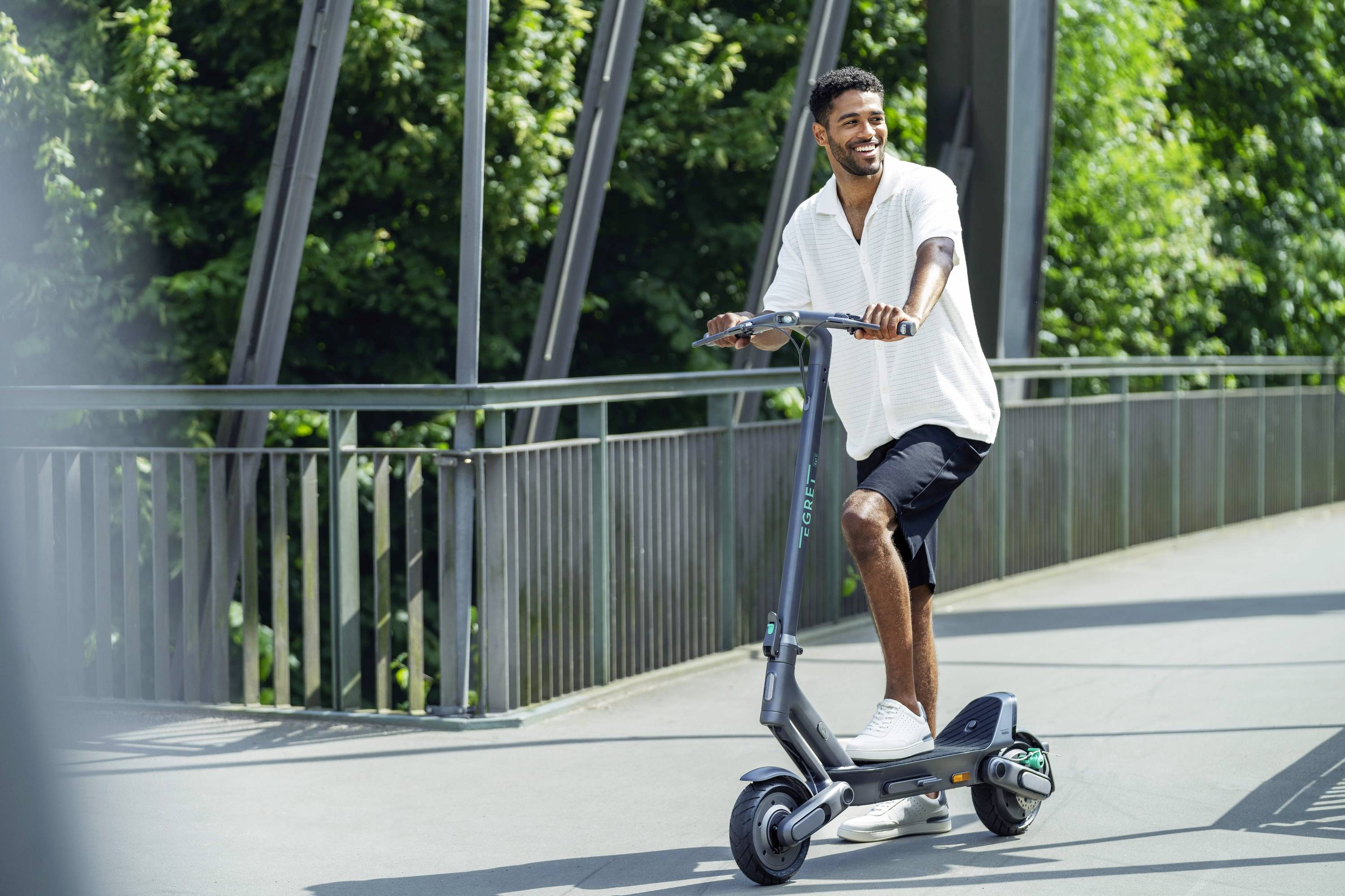 Ein Mann fährt einen E-Scooter auf einer Brücke an einem sonnigen Tag, umgeben von grüner Vegetation.