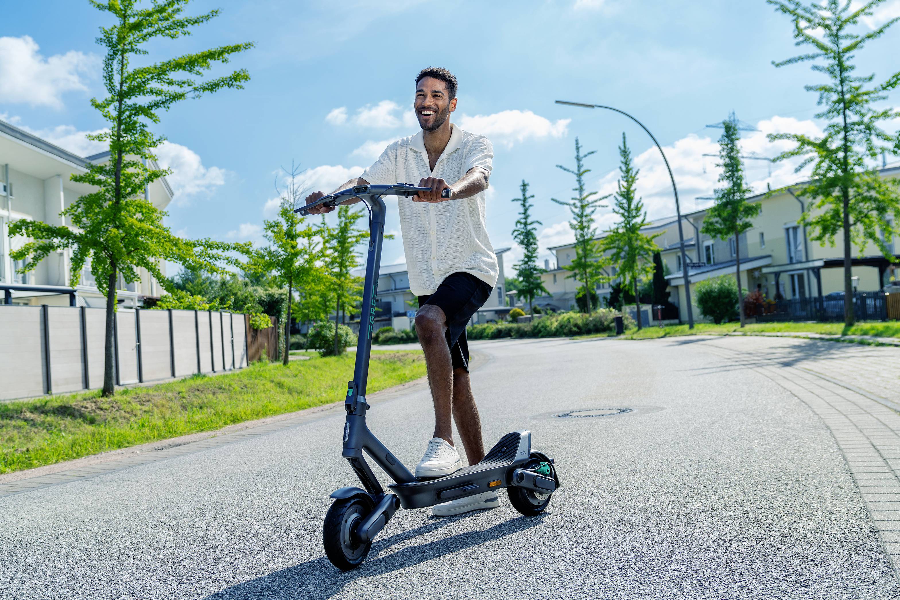 Ein Mann fährt bei sonnigem Wetter lächelnd einen Elektroroller auf einer ruhigen Wohnstraße entlang.