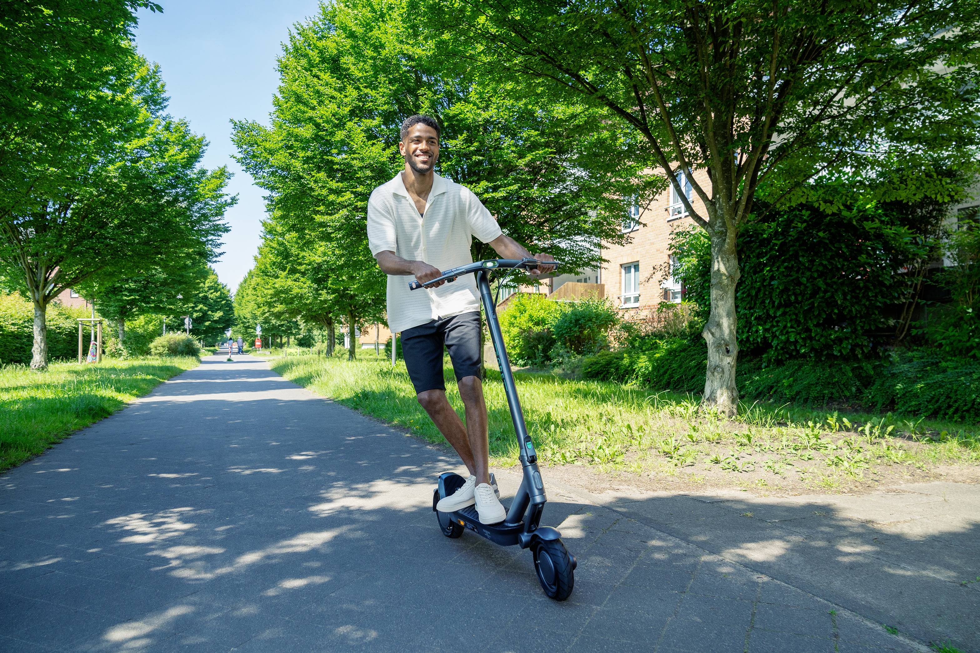 Ein Mann fährt fröhlich auf einem E-Scooter einen baumgesäumten Weg entlang. Die Sonne scheint und der Himmel ist blau.