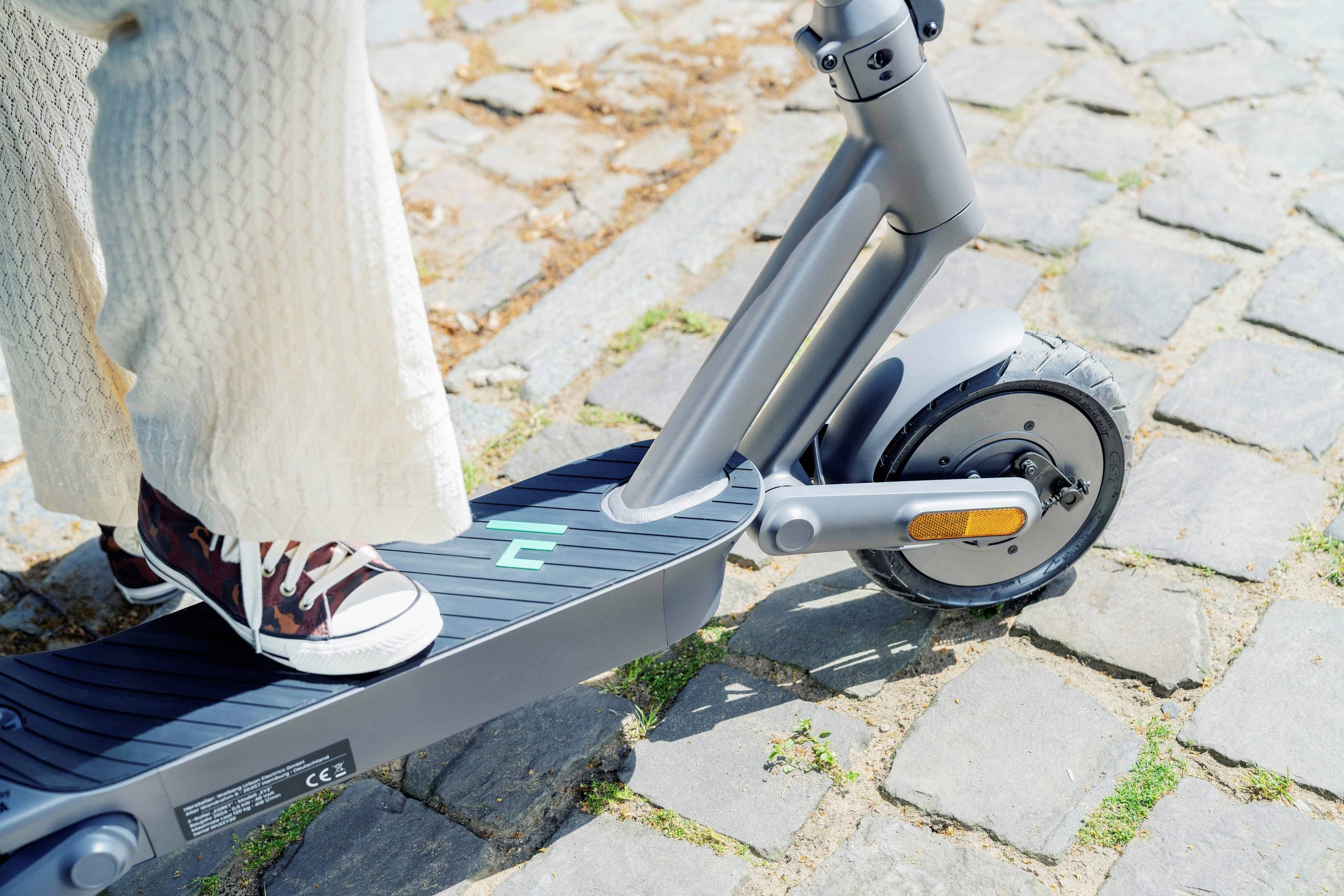 Person steht mit Converse-Schuhen auf einem Elektroroller, der auf einem gepflasterten Weg geparkt ist.