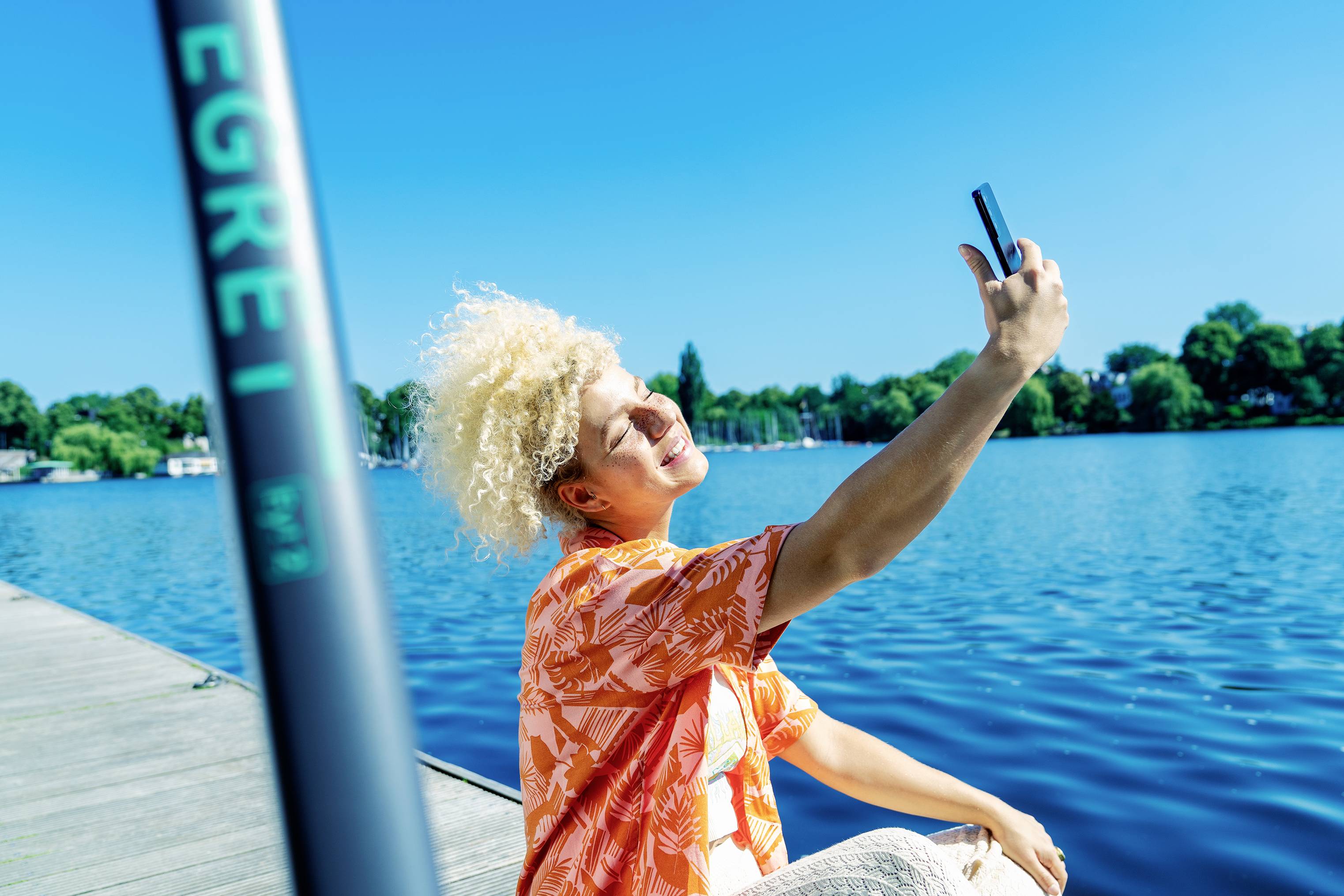 Eine Person fotografiert sich selbst mit einem Smartphone auf einem Steg am blauen See an einem sonnigen Tag.