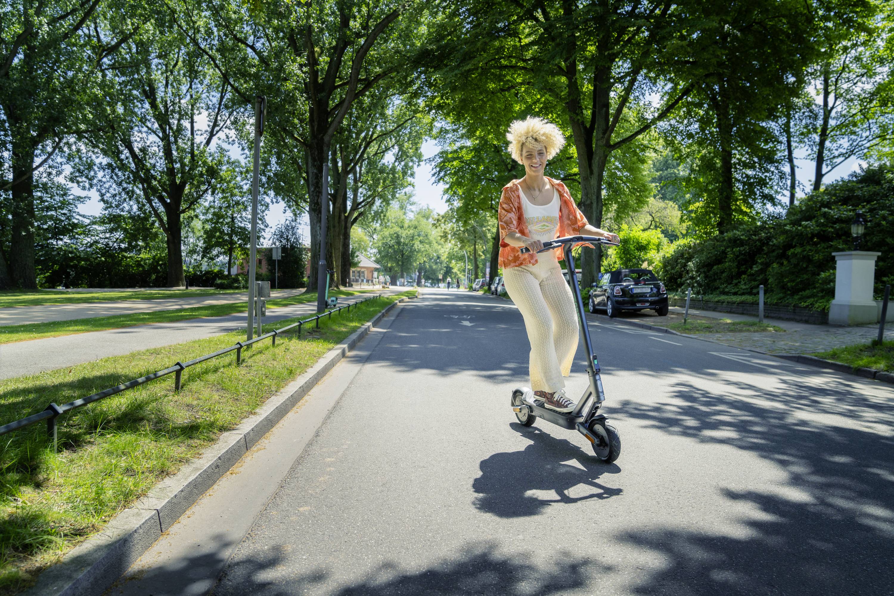 Eine Person fährt bei sonnigem Wetter einen Elektroroller auf einer von Bäumen gesäumten Straße.