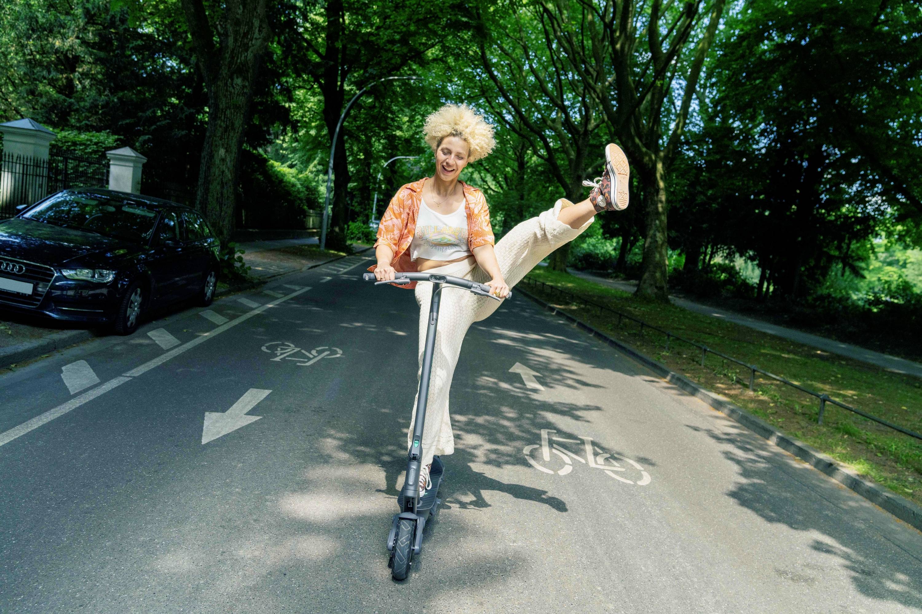 Eine Person mit lockigem Haar fährt fröhlich einen E-Scooter auf einer baumbestandenen Straße. Sie streckt ein Bein in die Luft.