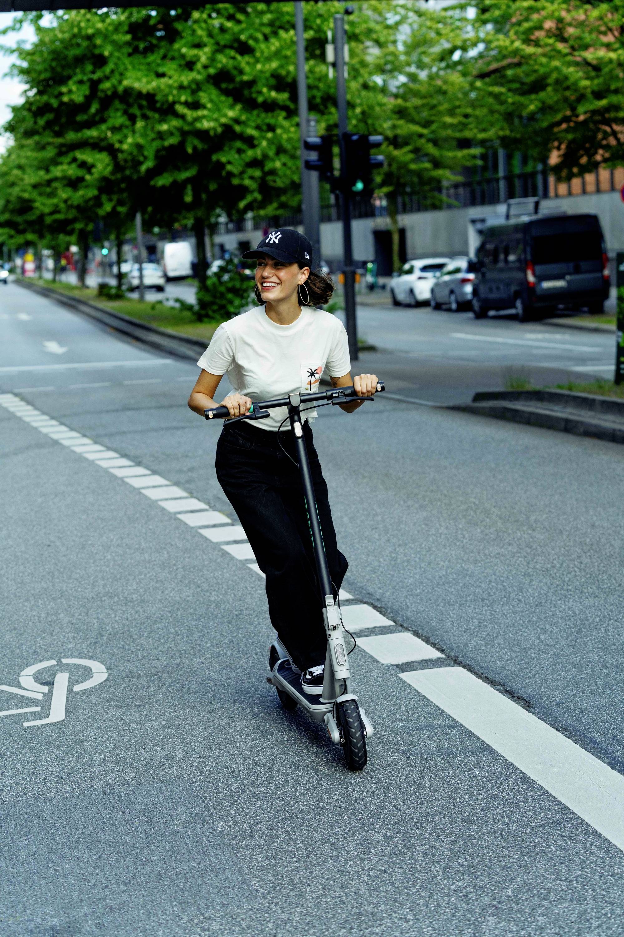 Eine Person fährt fröhlich mit einem E-Scooter auf einer städtischen Straße entlang einer markierten Spur. Bäume und Autos im Hintergrund.
